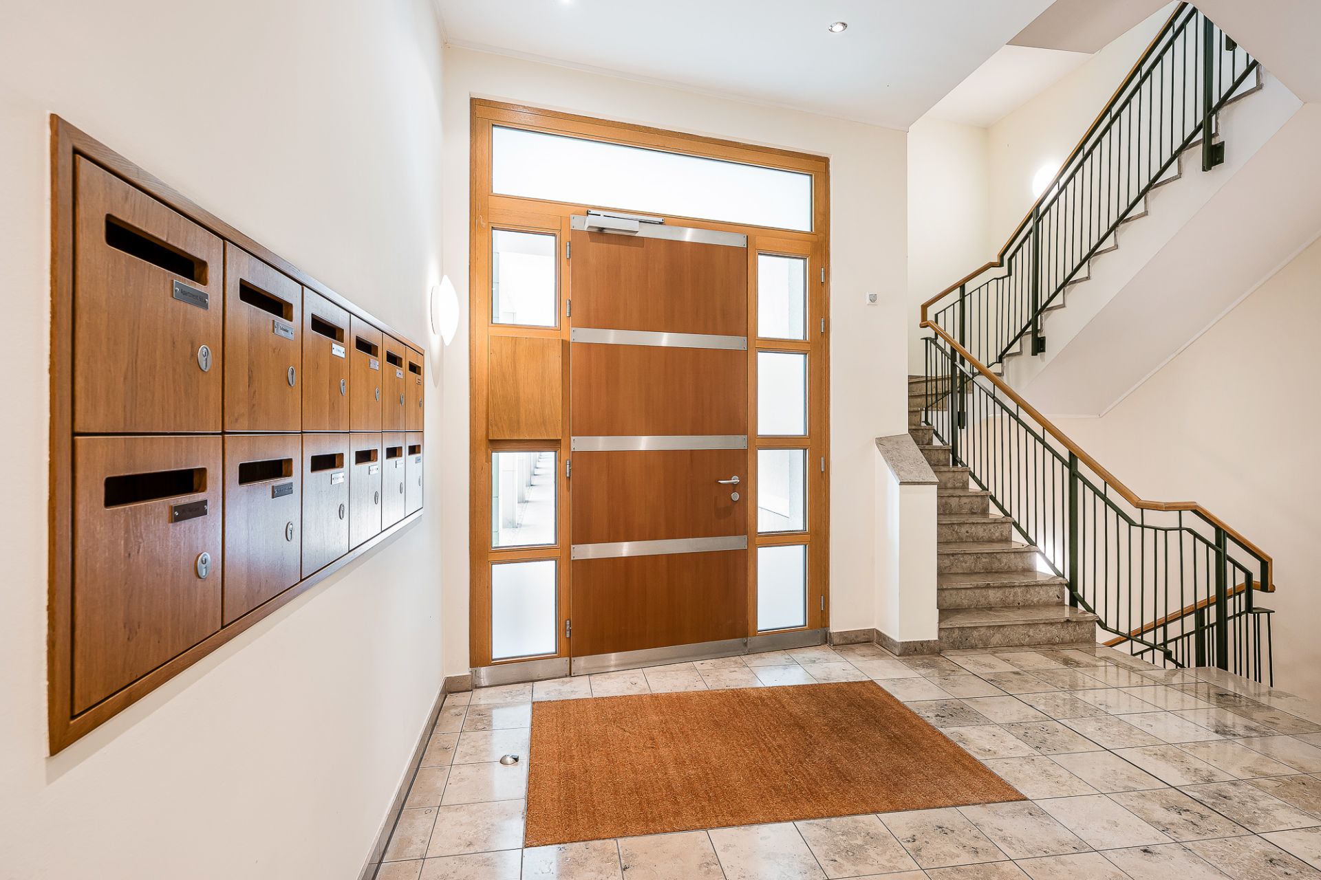 Heller Eingangsbereich mit Briefkästen, großer Holztür, Treppe und Fliesenboden.