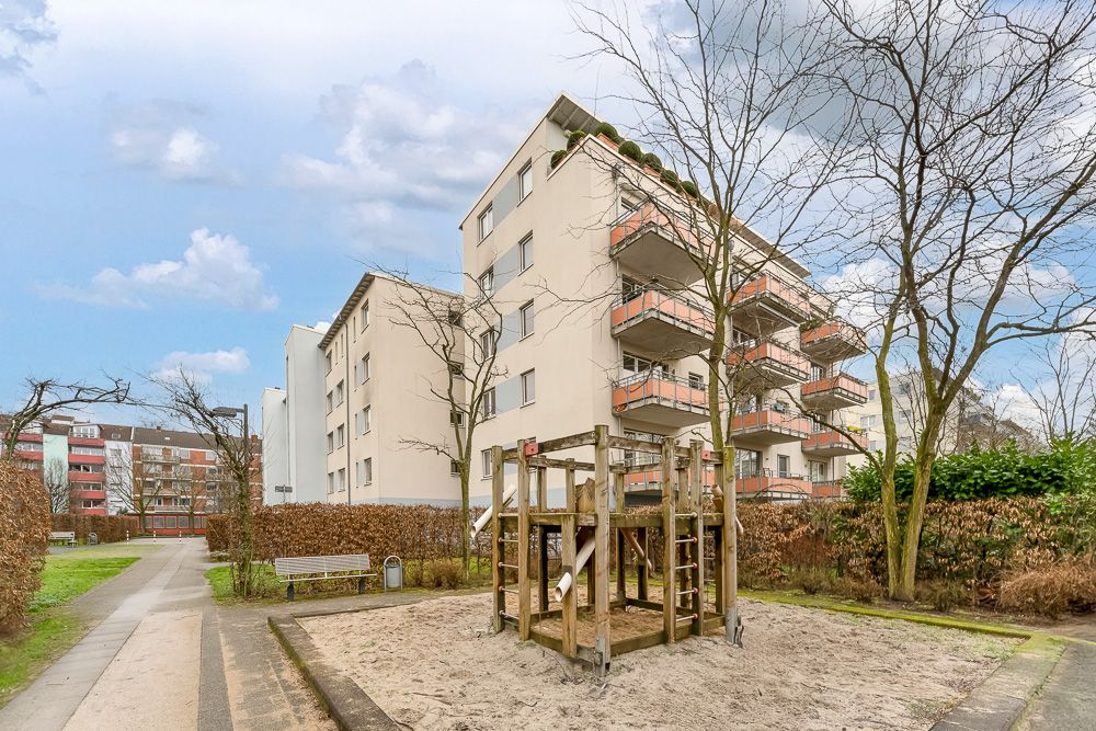 Mehrfamilienhaus mit Balkonen, davor ein Spielplatz mit Klettergerüst im Sand.