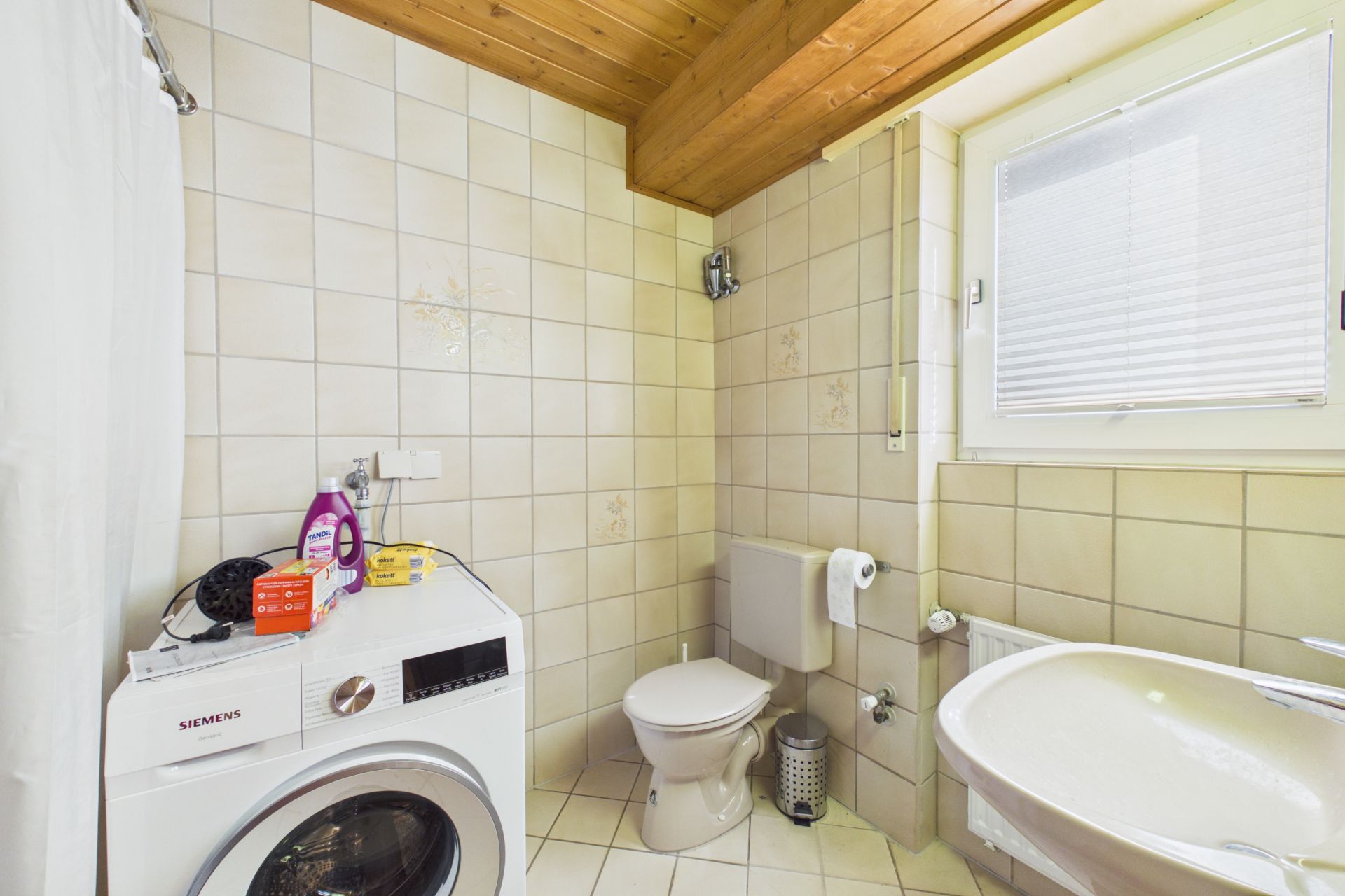 Helles Badezimmer mit Waschmaschine, Toilette, Waschbecken und Fenster mit Jalousie.