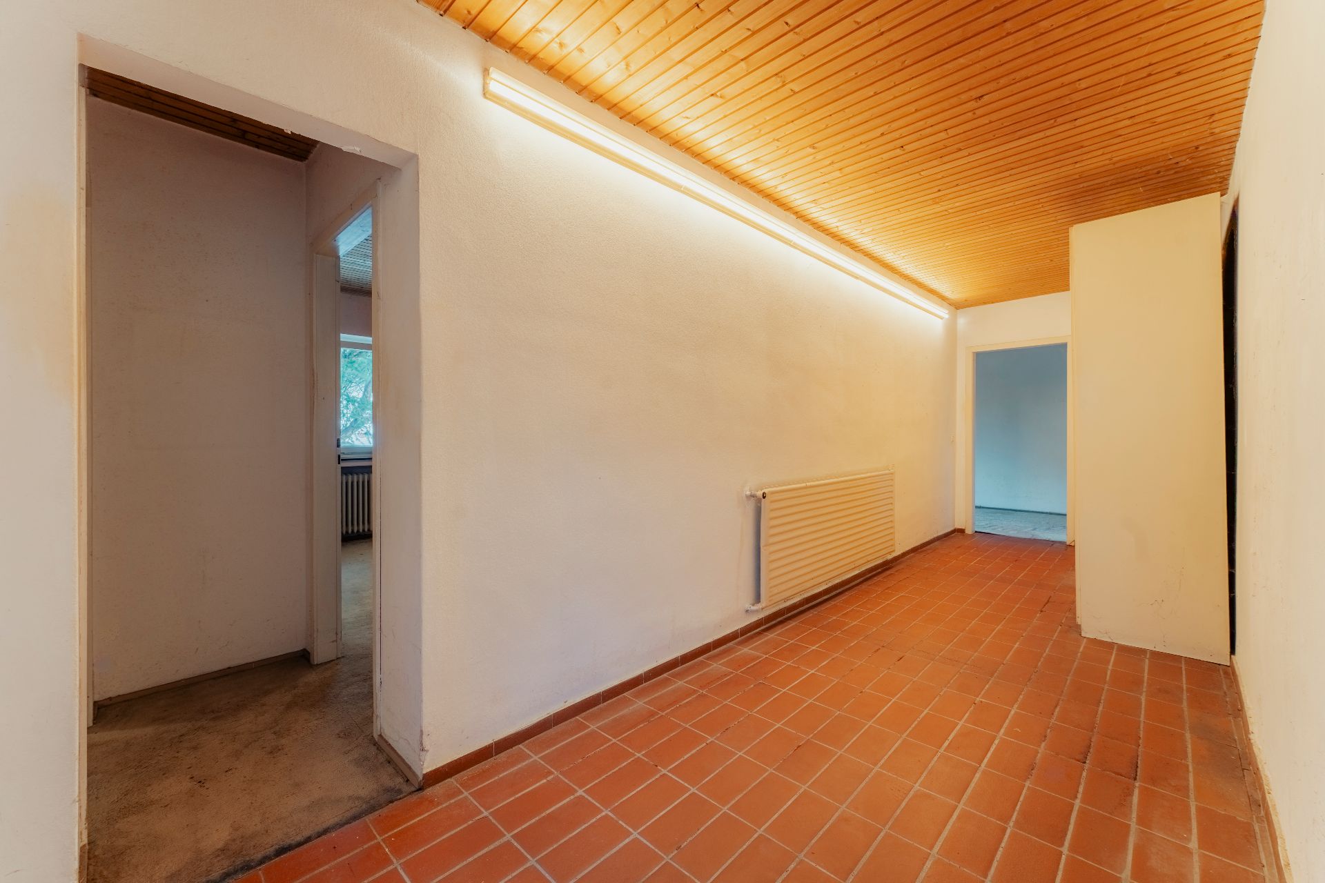 Flur mit terrakottafarbenen Fliesen, weißer Wand und Holzdecke, Türen zu angrenzenden Zimmern.