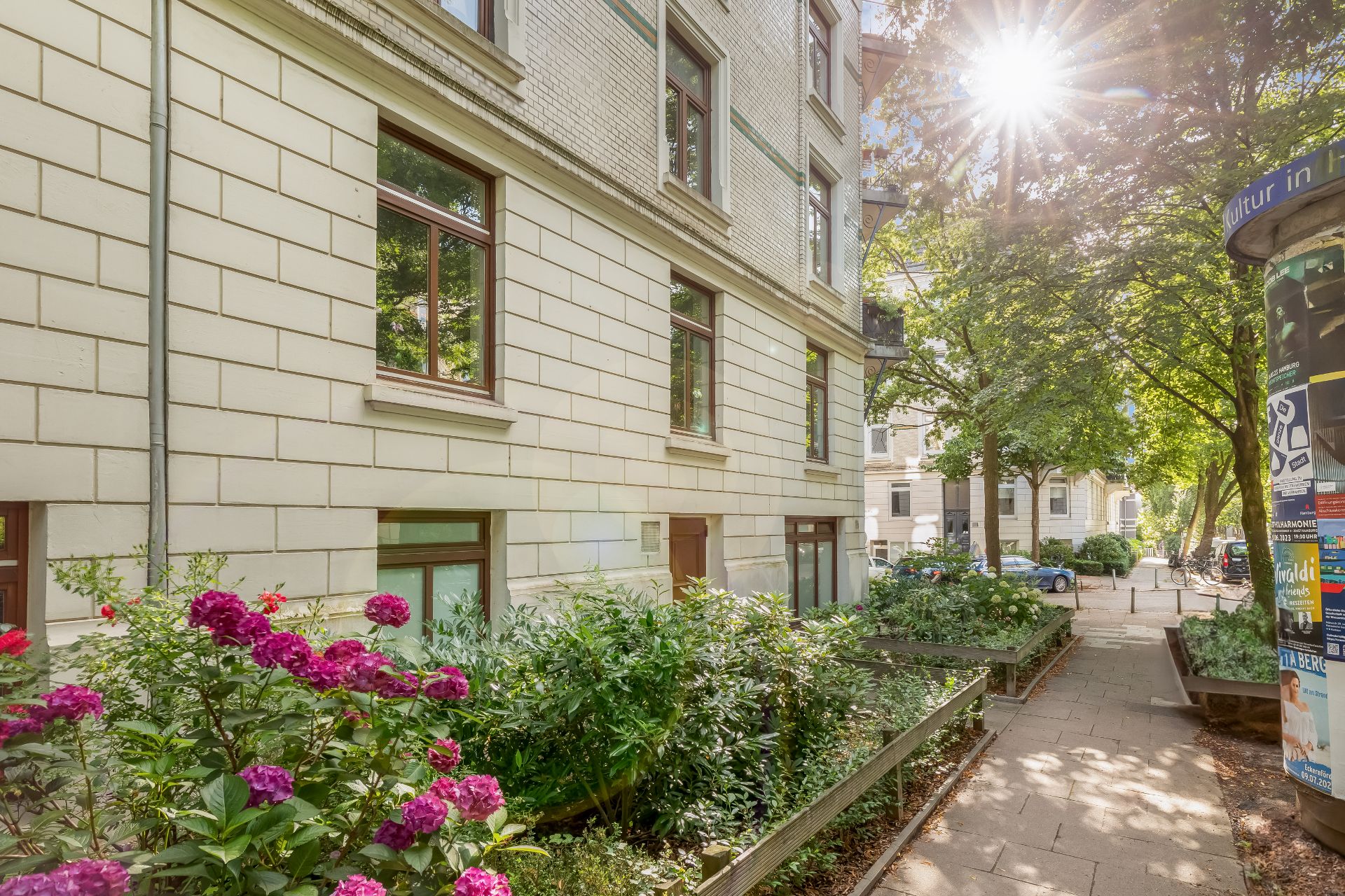 Wohnhaus mit hellem Mauerwerk, Blumenbeeten und Gehweg im Vordergrund bei Sonnenschein.
