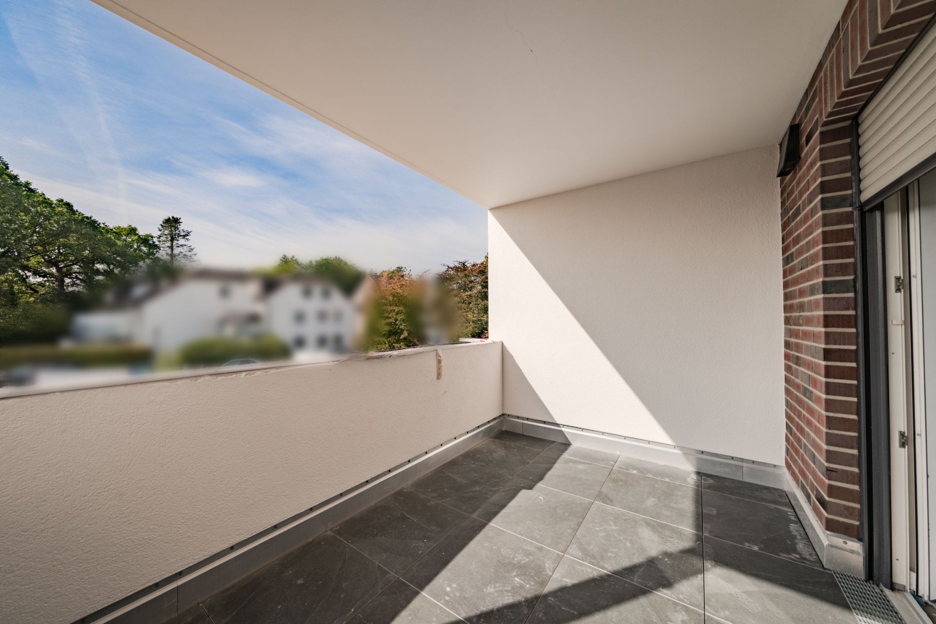 Überdachter Balkon mit Fliesenboden und Blick ins Grüne, seitlich Backsteinwand.