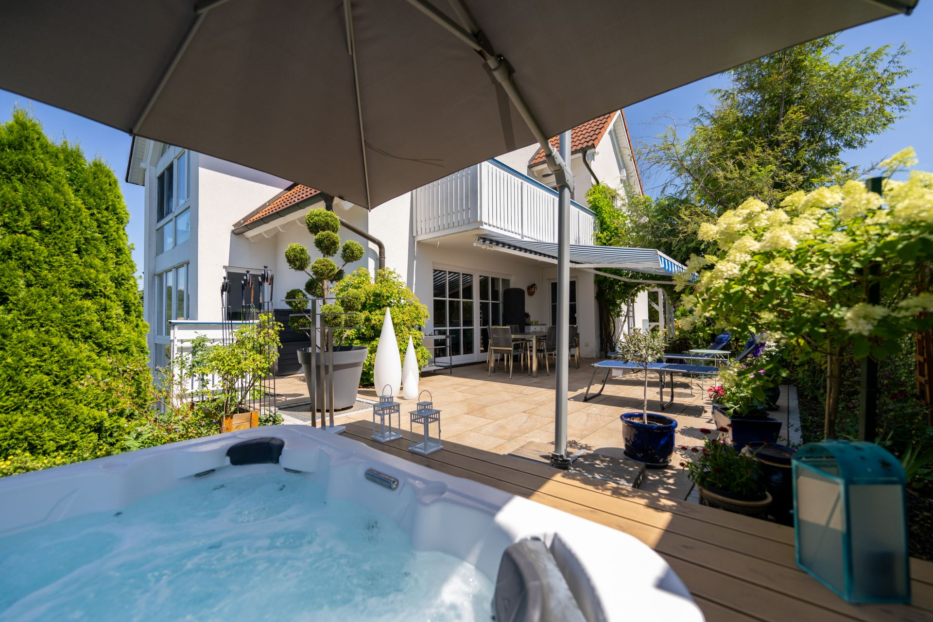 Garten mit Whirlpool, Terrasse, Sonnenschirm und Blumen neben einem Wohnhaus bei Sonnenschein.