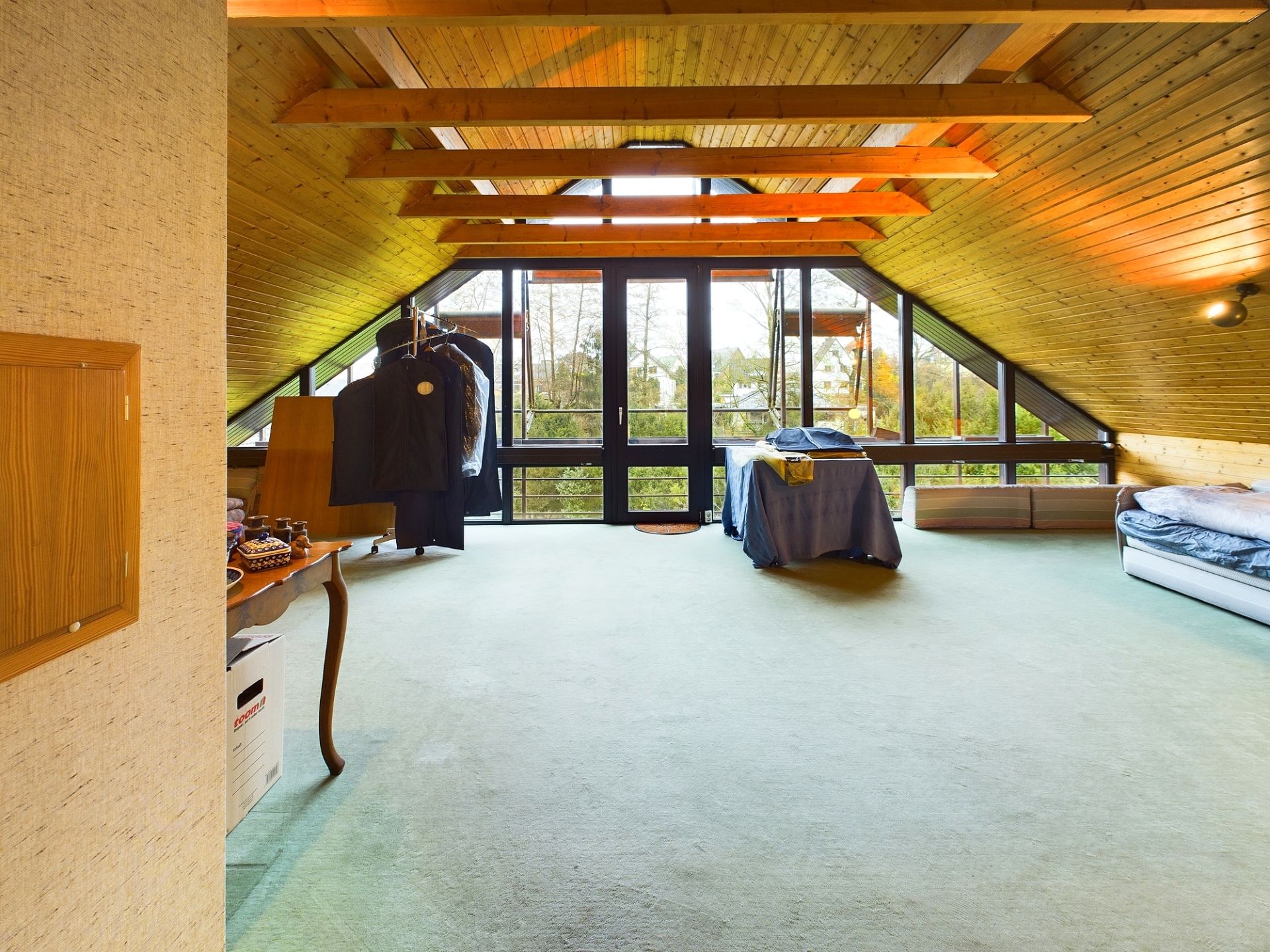 Dachzimmer mit schrägen Holzdecken, großem Fenster, Bett und Tisch.