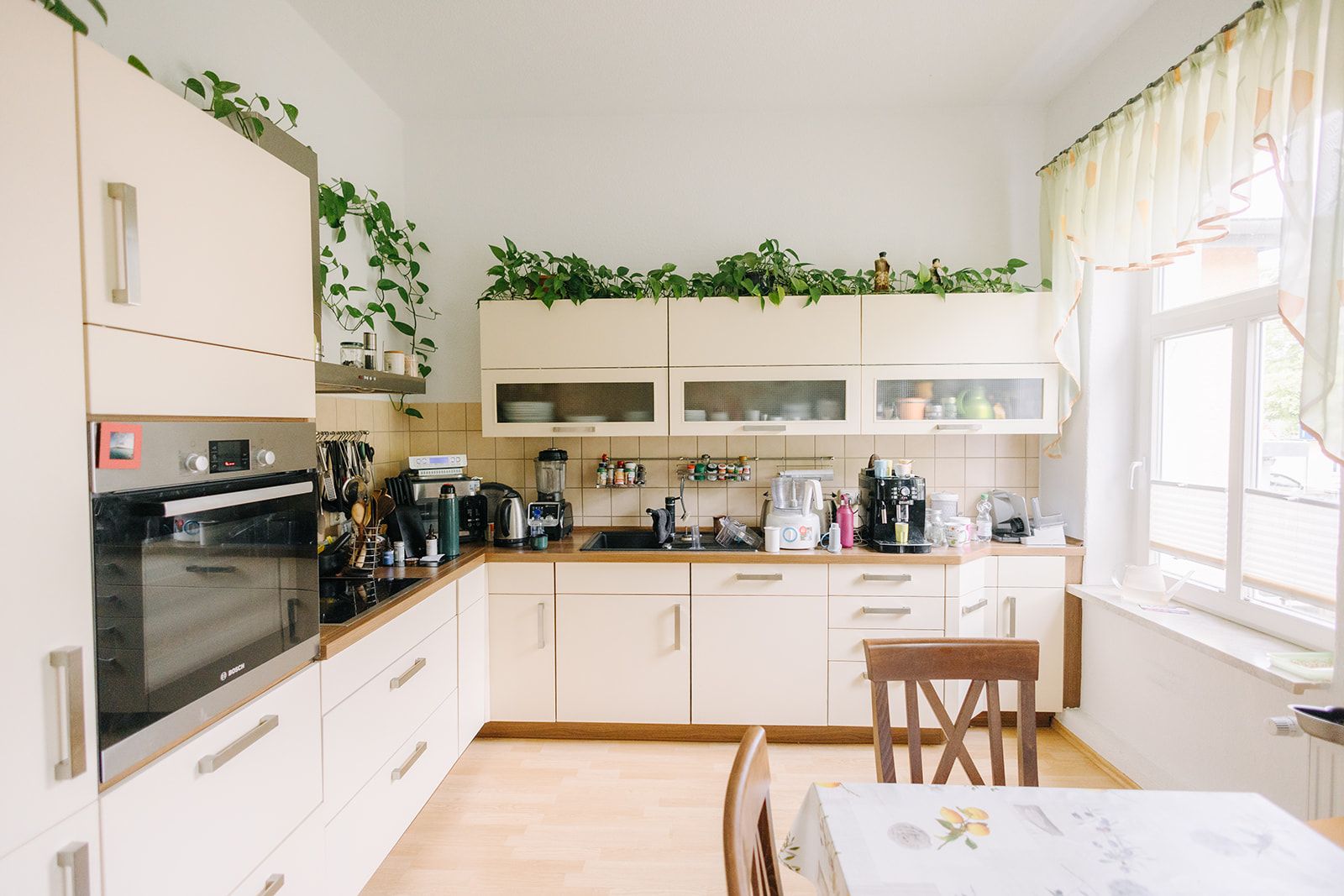 Helle Küche mit weißen Schränken, Geräten, Pflanzen und großem Fenster, Esstisch im Vordergrund.