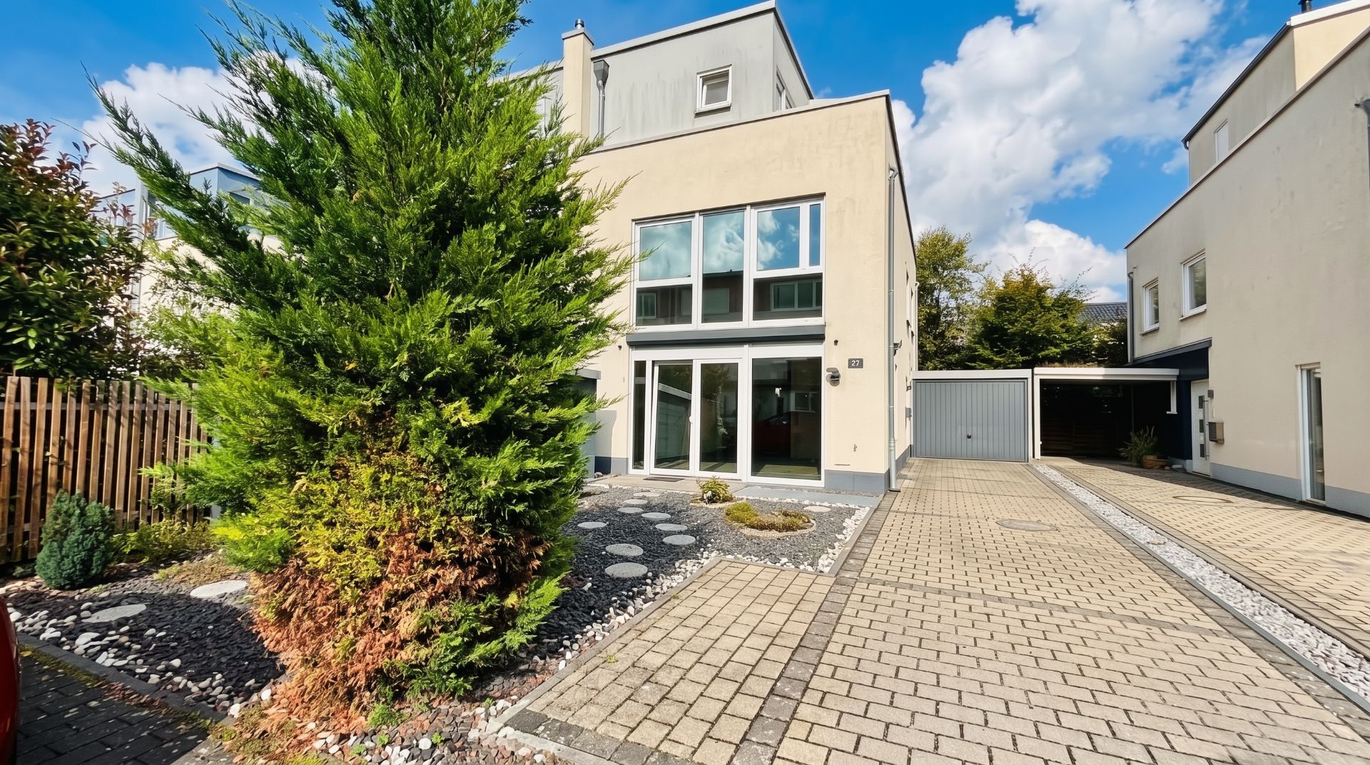 Modernes Wohnhaus mit großen Fenstern, gepflasterter Einfahrt und Vorgarten mit Baum.