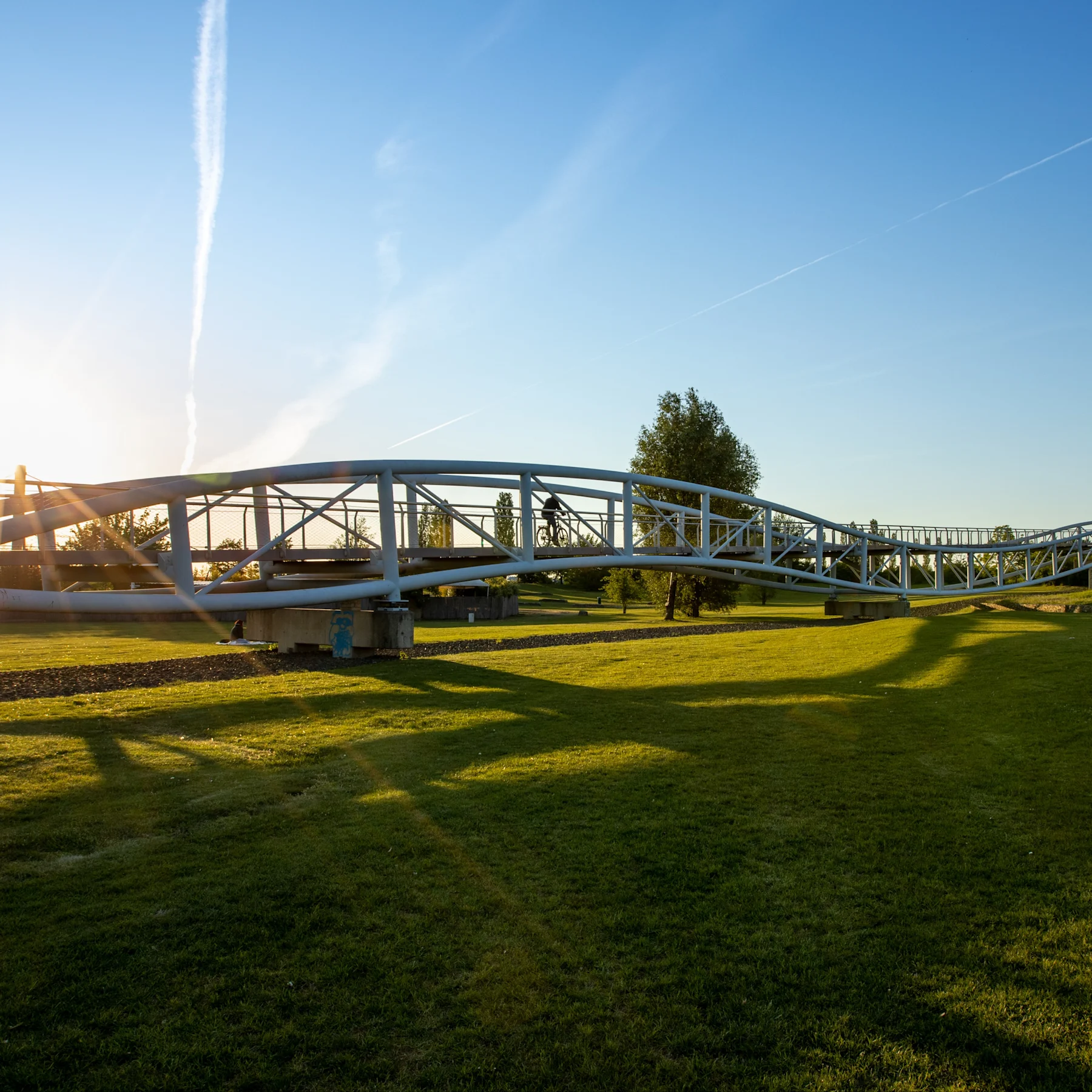 Metallbrücke über einer Wiese bei Sonnenuntergang, mit Bäumen im Hintergrund.