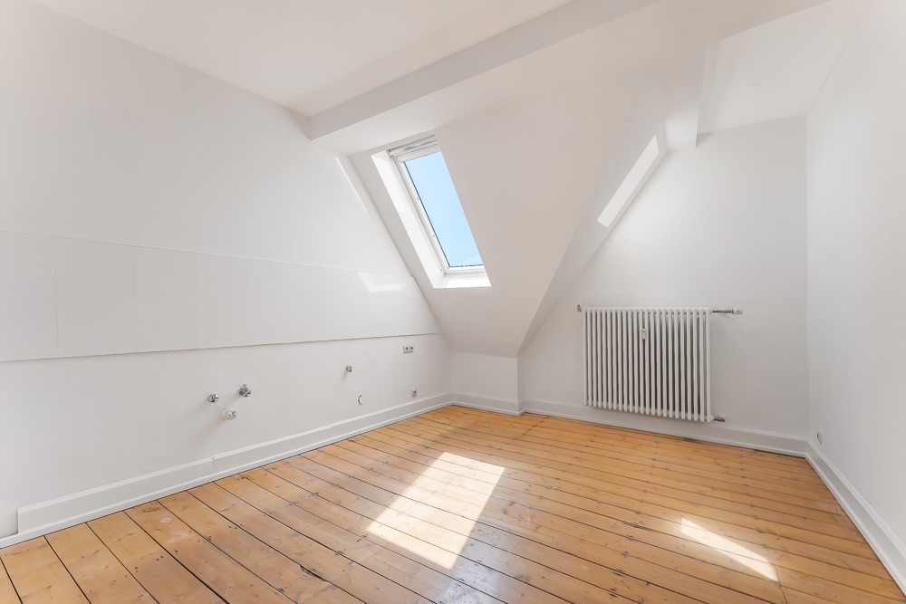 Heller, leerer Raum mit Holzfußboden, Dachfenster und Heizkörper an der Wand.
