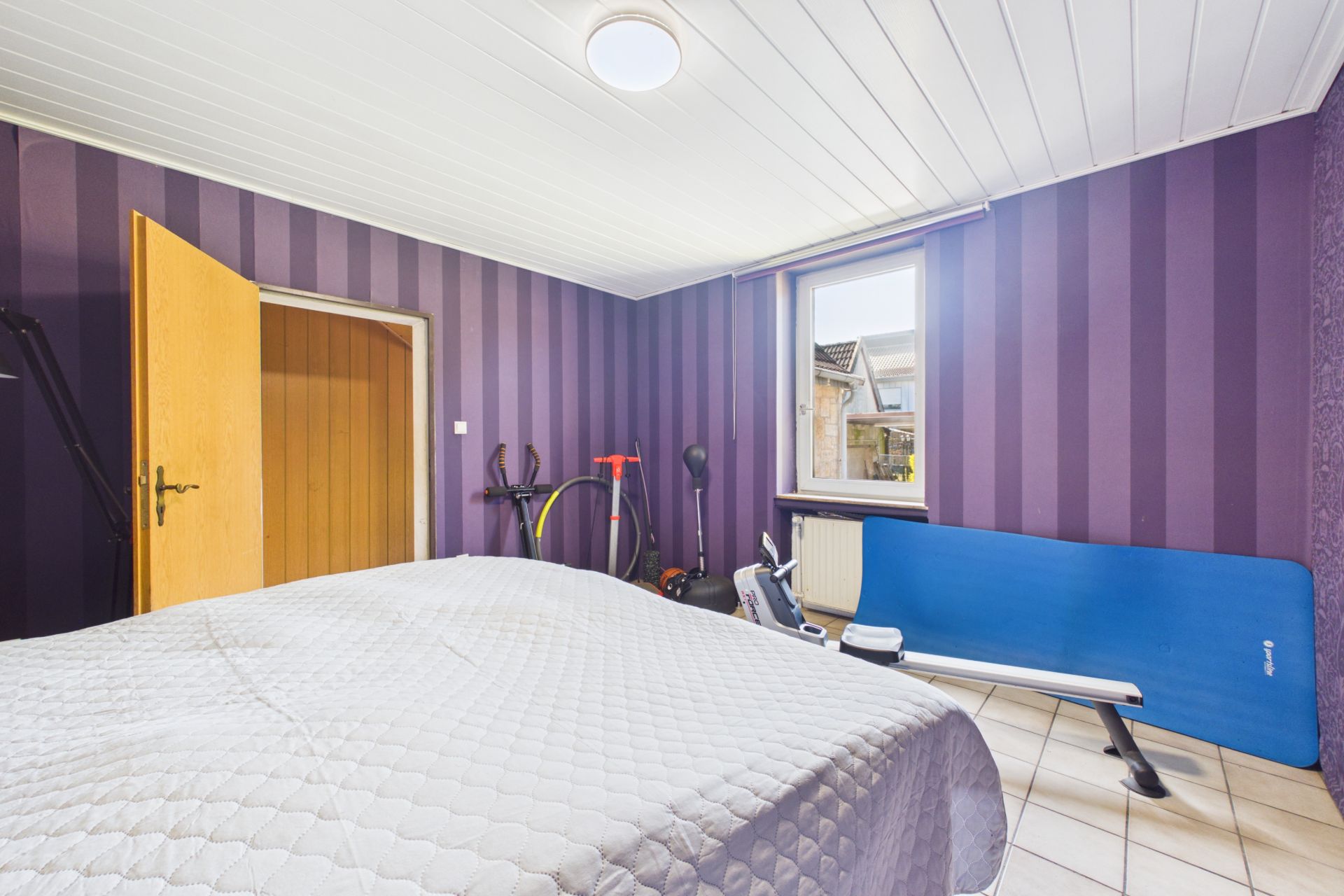 Schlafzimmer mit lila gestreifter Wand, Bett, offenem Holztür, Fitnessgerät und Fahrrad.