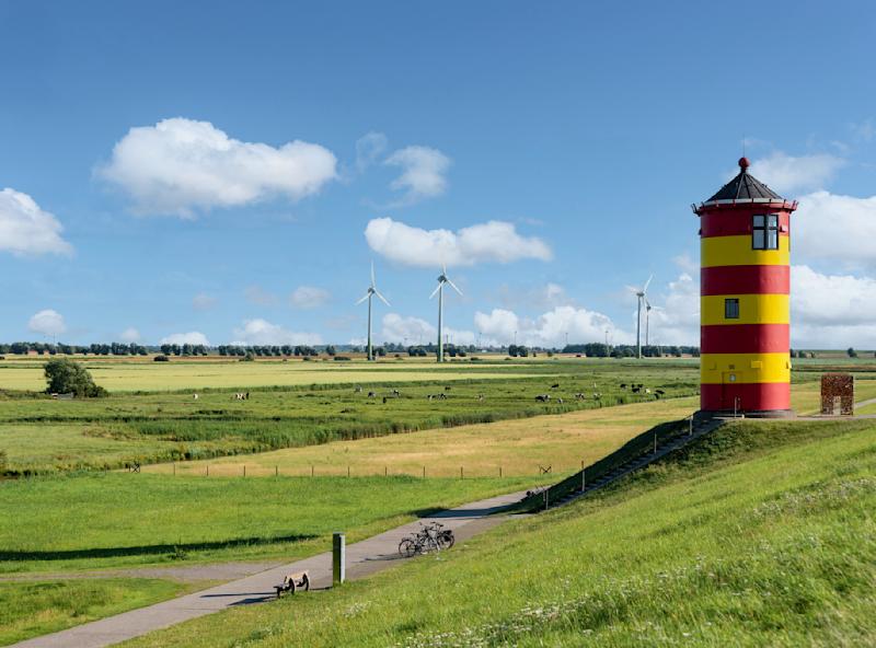Roter und gelber Turm auf einem Deich mit Blick auf grüne Felder und Windräder im Hintergrund.
