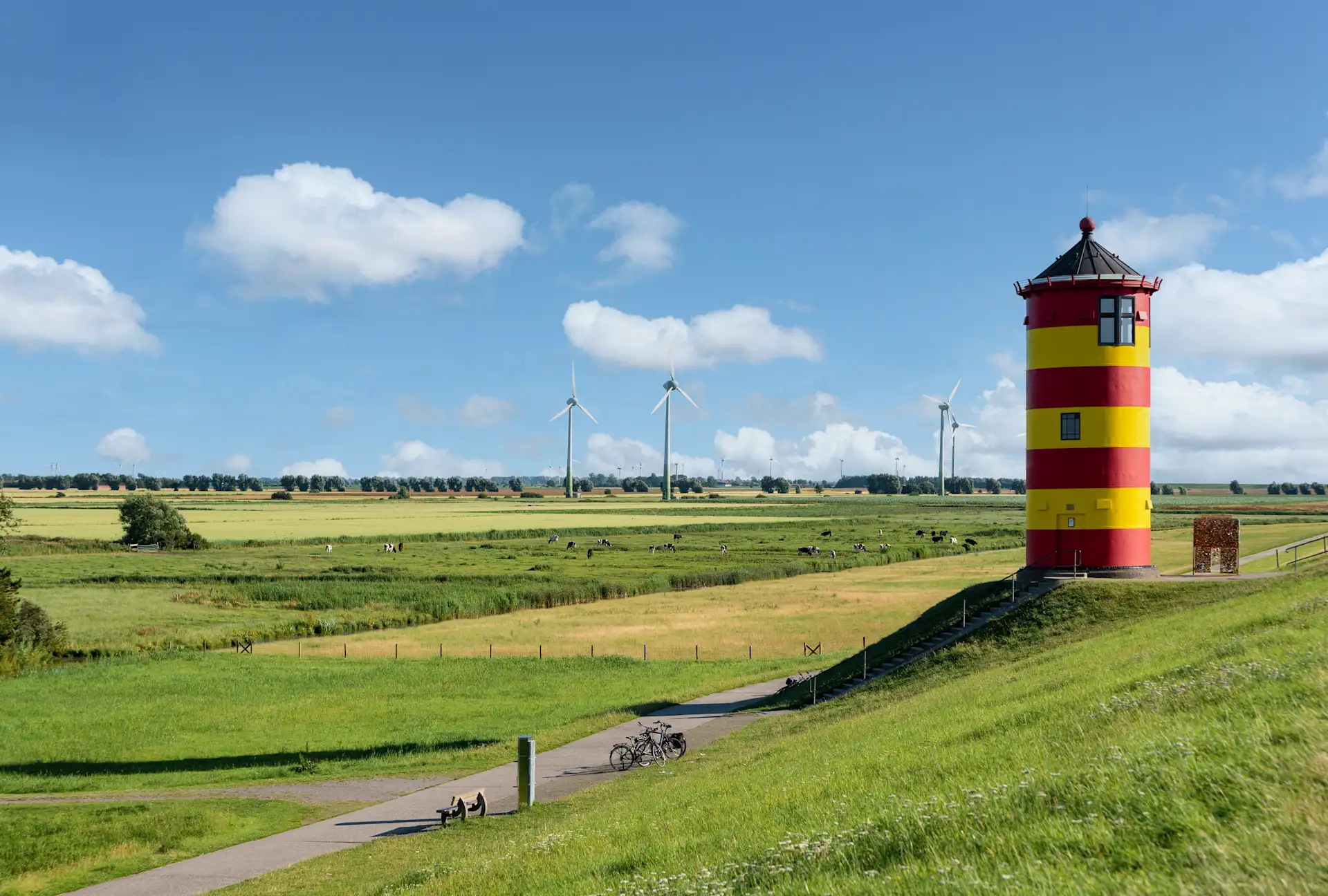 Roter und gelber Turm auf einem Deich mit Blick auf grüne Felder und Windräder im Hintergrund.