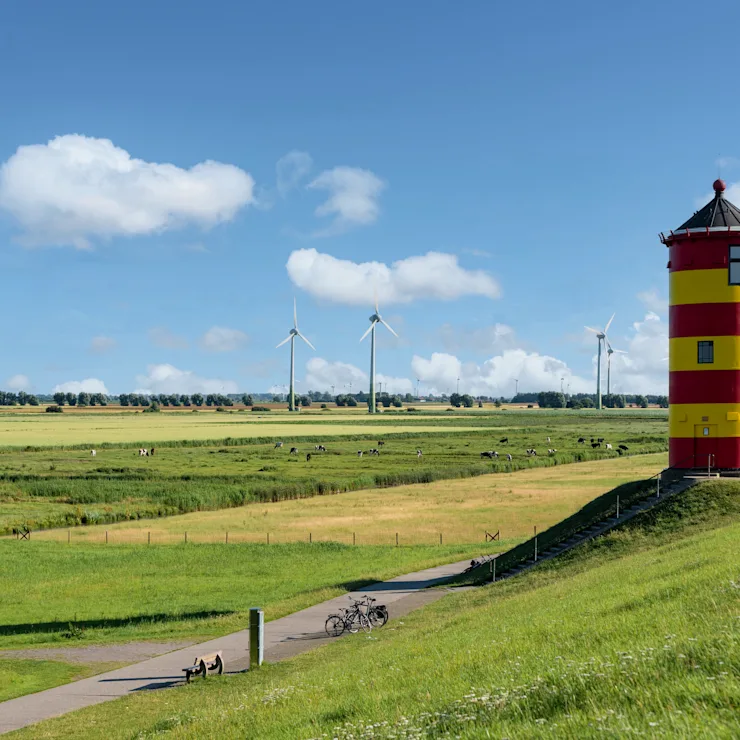 Roter und gelber Turm auf einem Deich mit Blick auf grüne Felder und Windräder im Hintergrund.