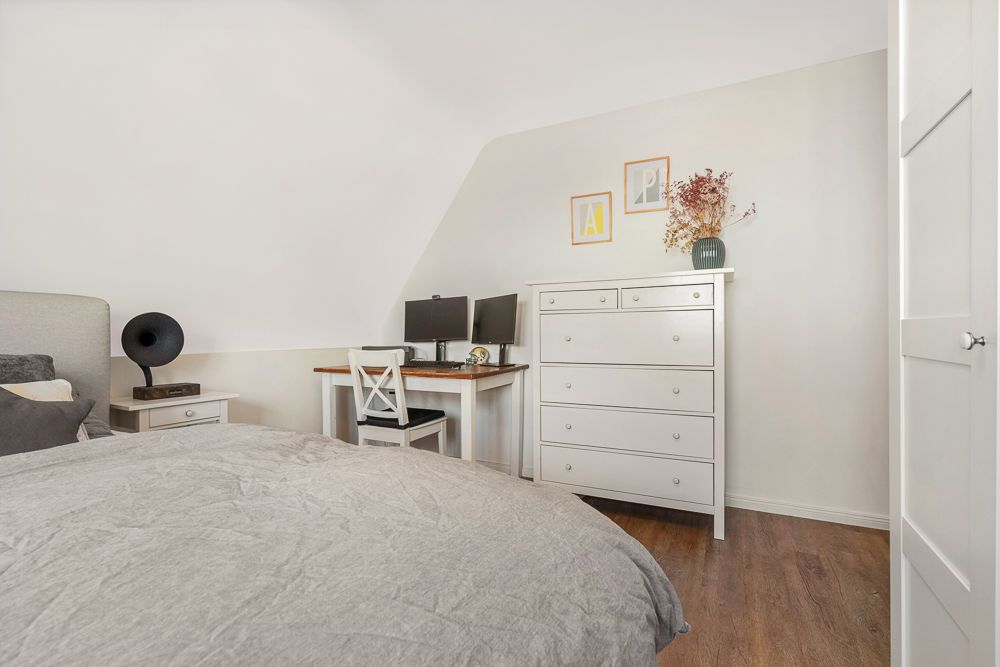 Helles Schlafzimmer mit Schreibtisch, zwei Monitoren, Kommode und Dekoration auf Holzfußboden.