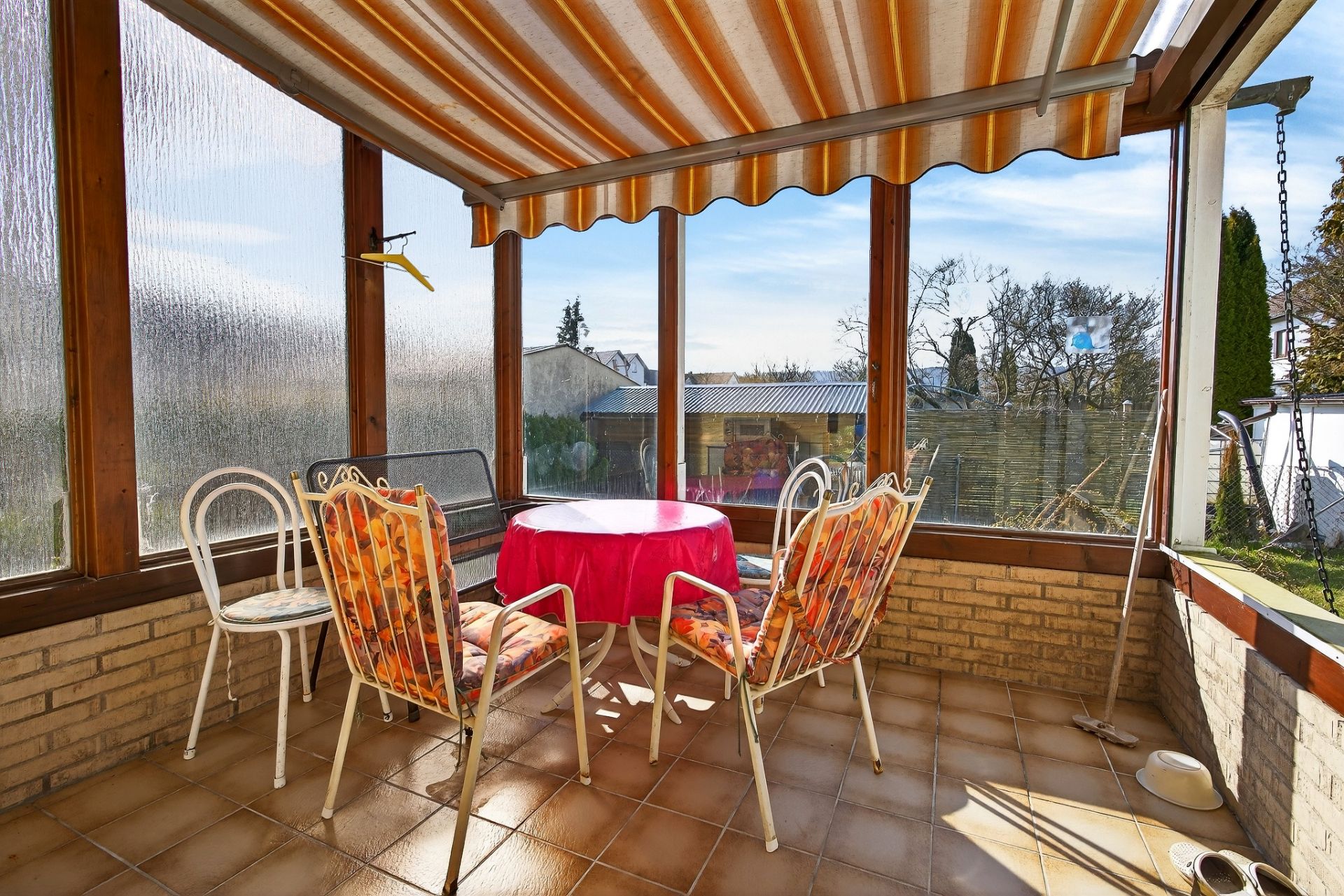 Wintergarten mit Terrakottafliesen, Tisch mit rotem Tuch und vier Stühlen, Ausblick auf Garten.