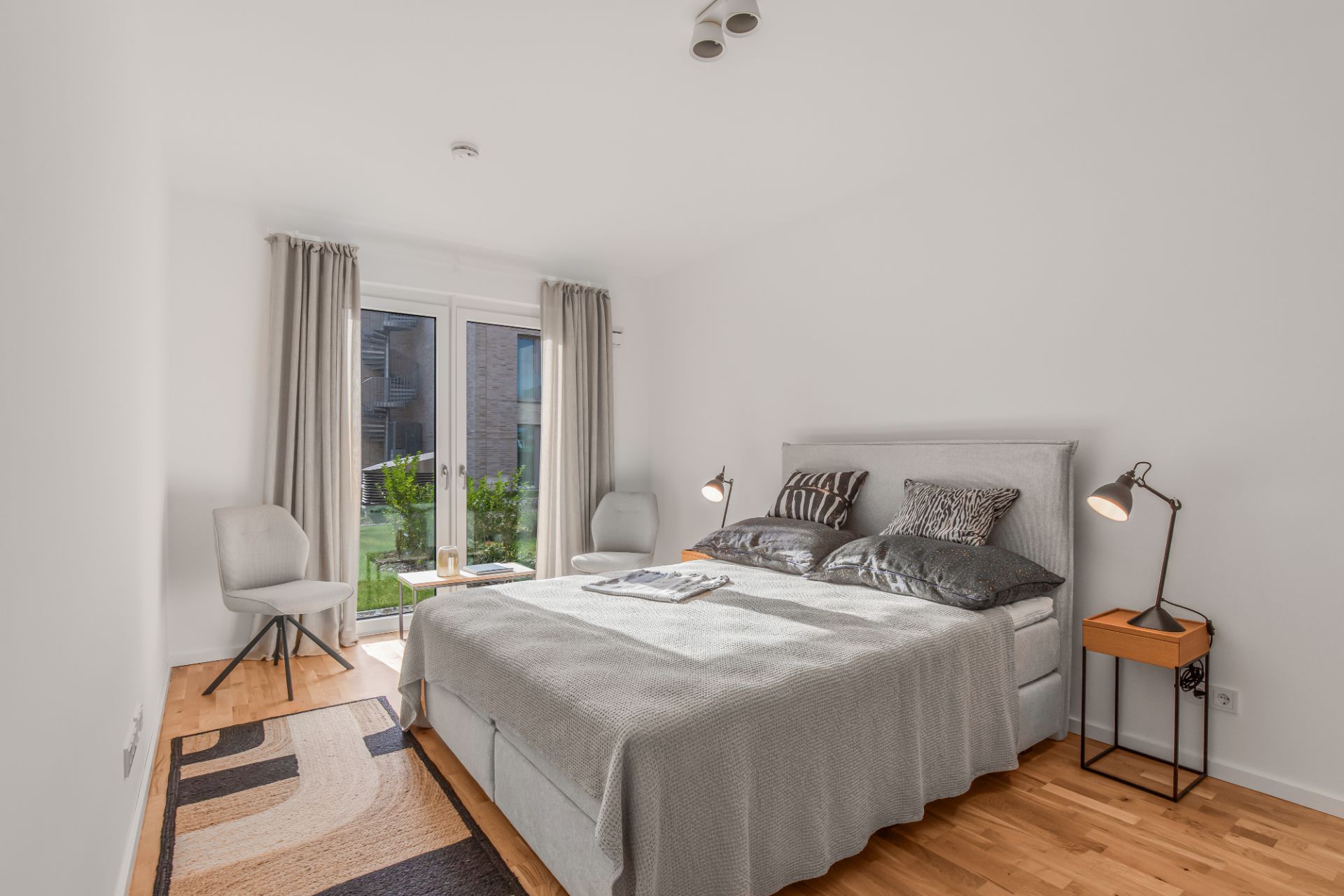 Helles Schlafzimmer mit Doppelbett, zwei Nachttischen, zwei Stühlen und Fenster mit Blick nach draußen.