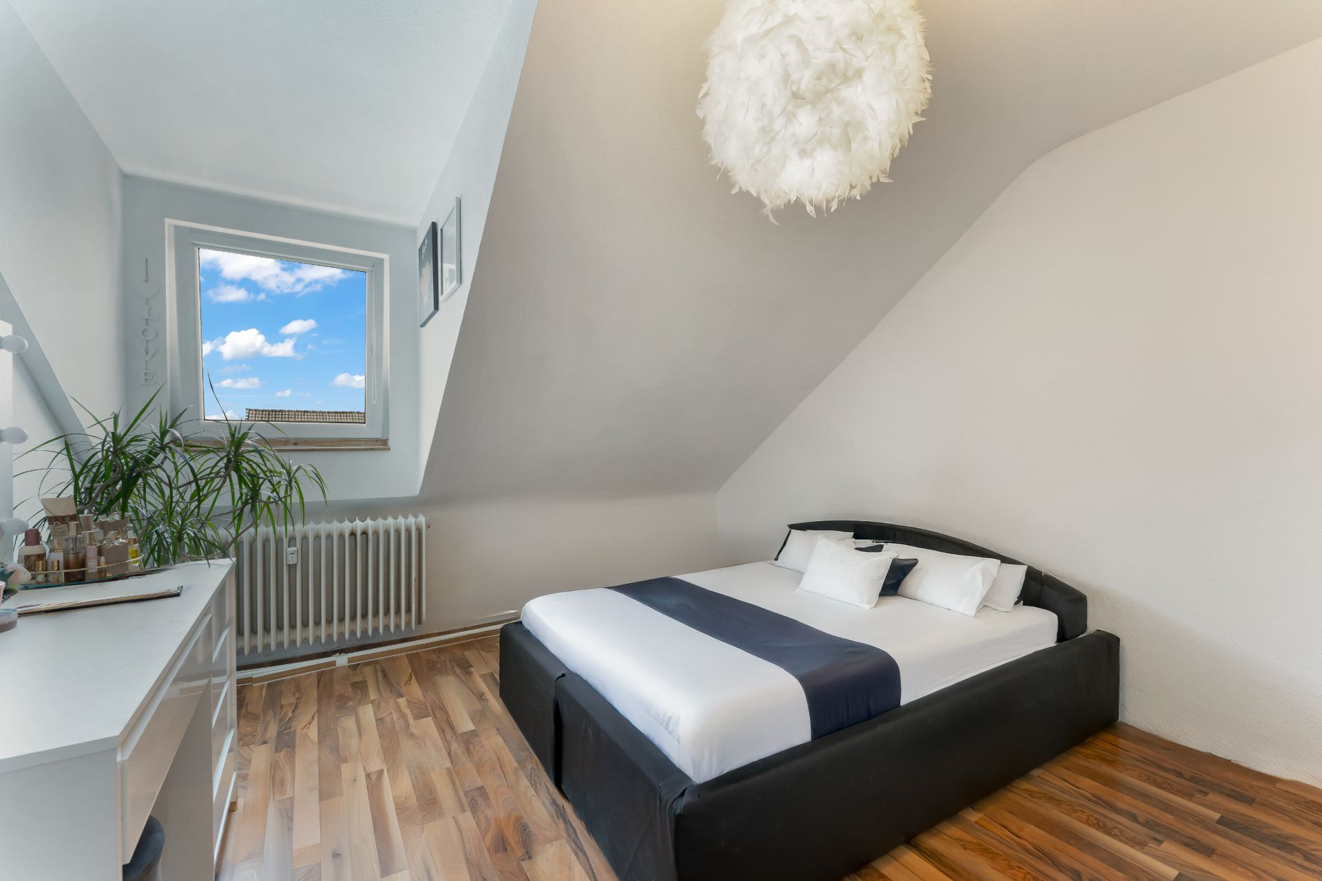 Schlafzimmer mit Doppelbett unter Dachschräge, Kommode, Pflanze und Fenster mit Blick nach draußen.