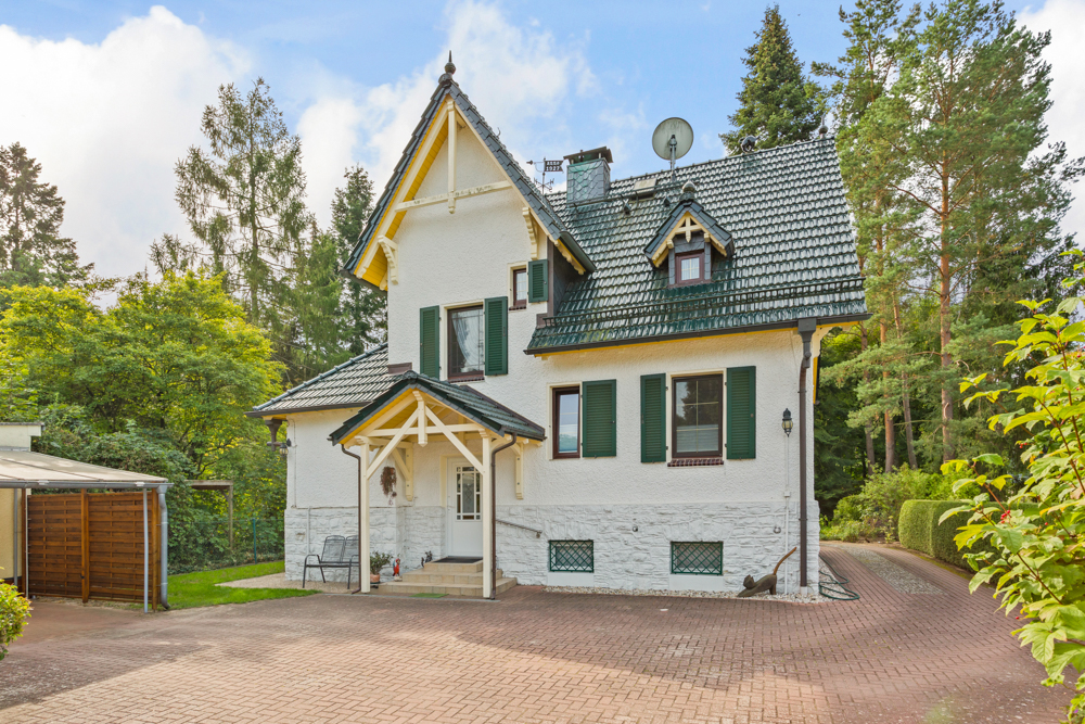 Weißes Einfamilienhaus mit grünem Dach, Fensterläden und Auffahrt, umgeben von Bäumen.
