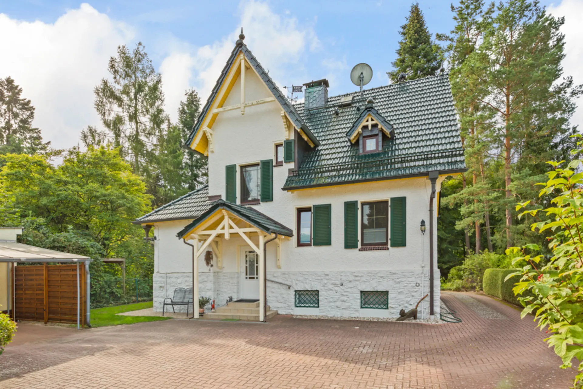 Weißes Einfamilienhaus mit grünem Dach, Fensterläden und Auffahrt, umgeben von Bäumen.