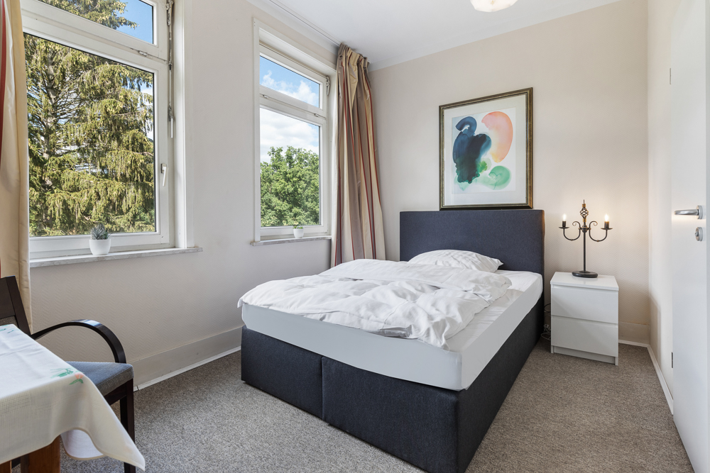 Helles Schlafzimmer mit Doppelbett, zwei Fenstern, Nachttisch und Wandbild.