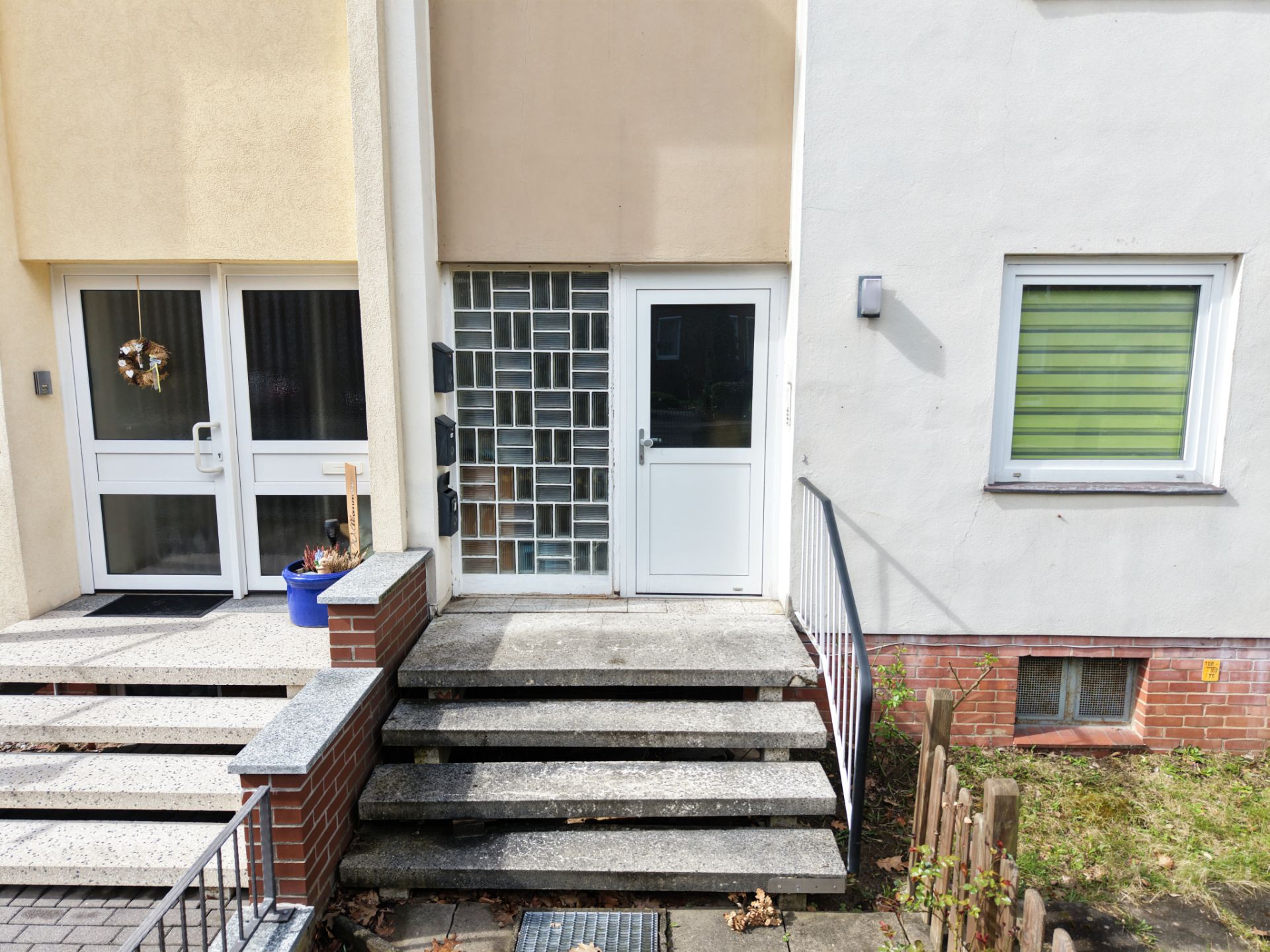Eingangsbereich eines Wohnhauses mit Treppen, zwei Türen und einem Fenster mit grünem Rollladen.