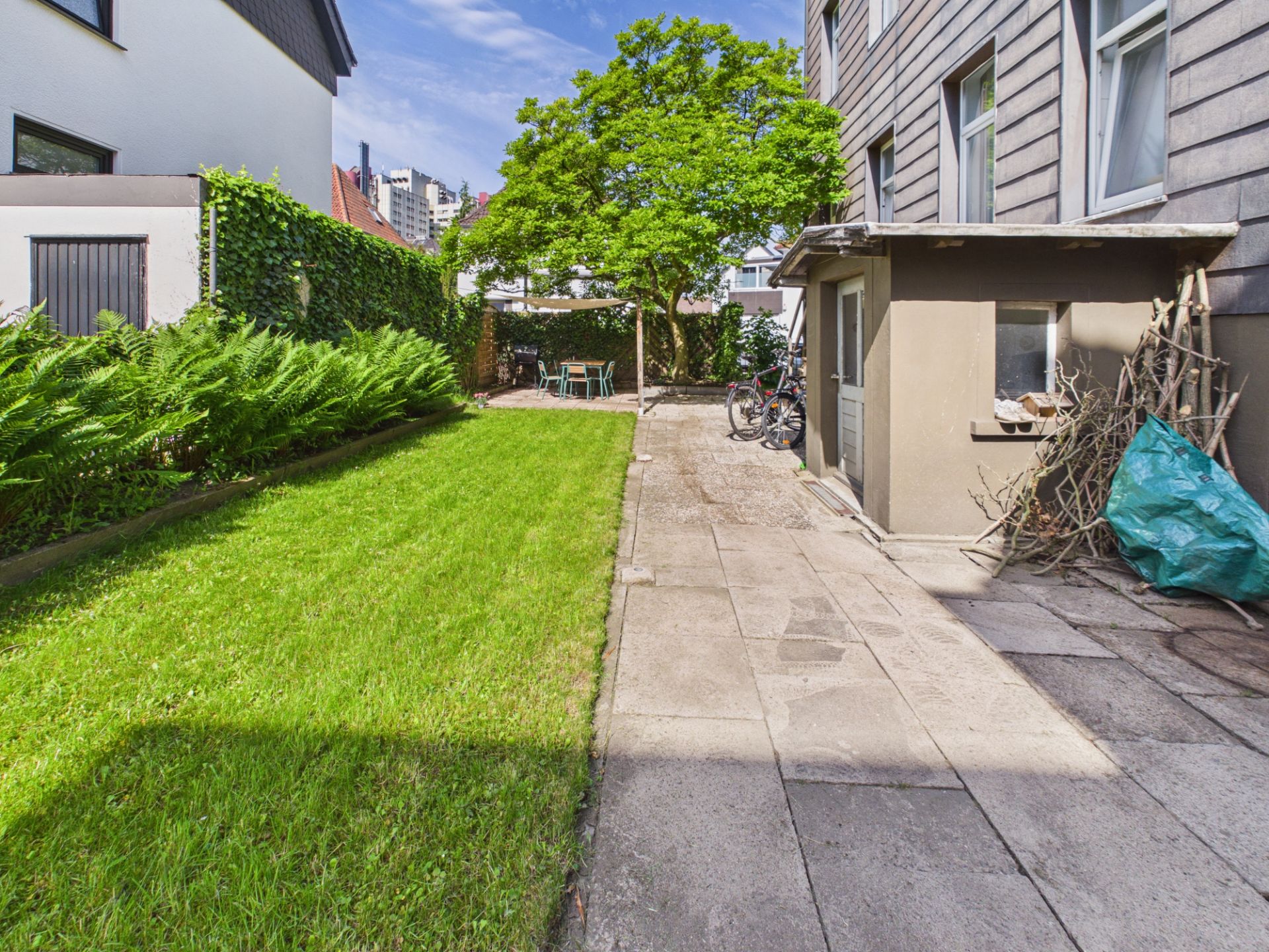 Garten mit Rasenfläche, Gehweg, Bäumen und einem Sitzbereich neben einem Wohnhaus.