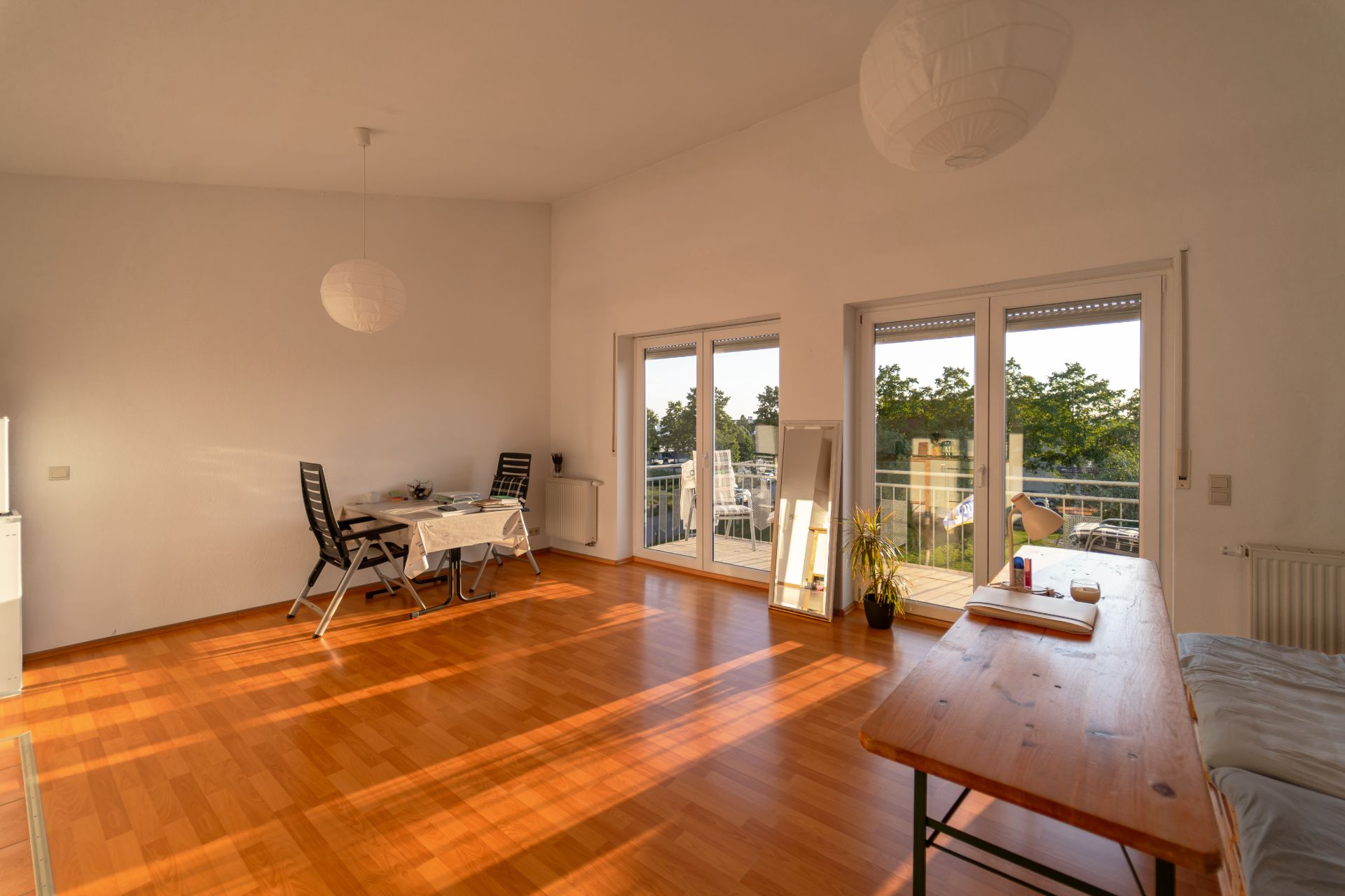 Helles Wohnzimmer mit Holzboden, Esstisch, Schreibtisch und Zugang zu einem Balkon mit Glas­türen.