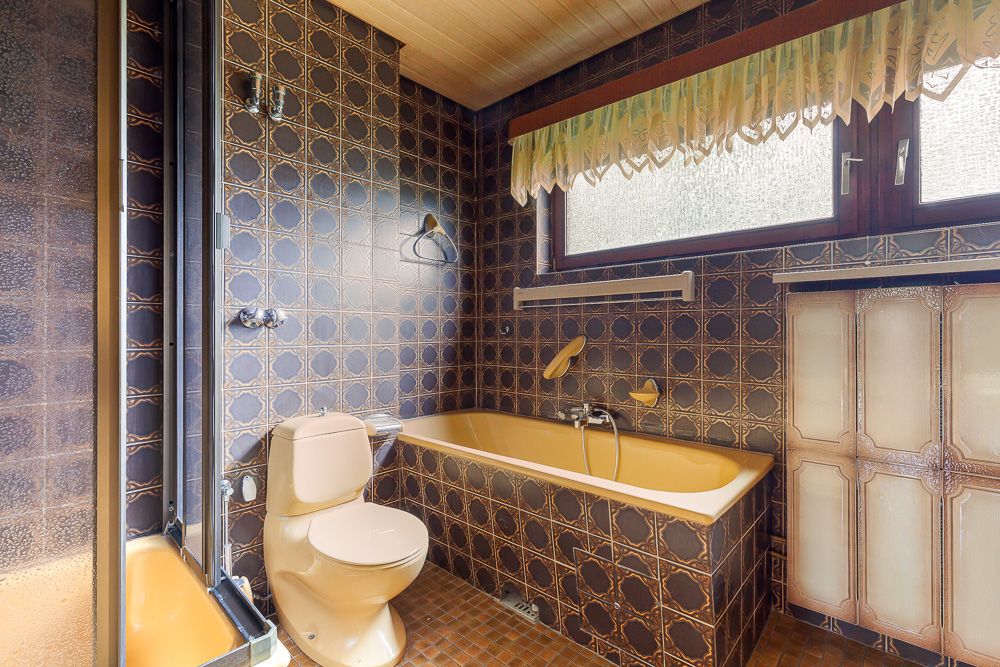 Badezimmer mit braunen Fliesen, gelber Badewanne und Toilette, Fenster mit Vorhang.