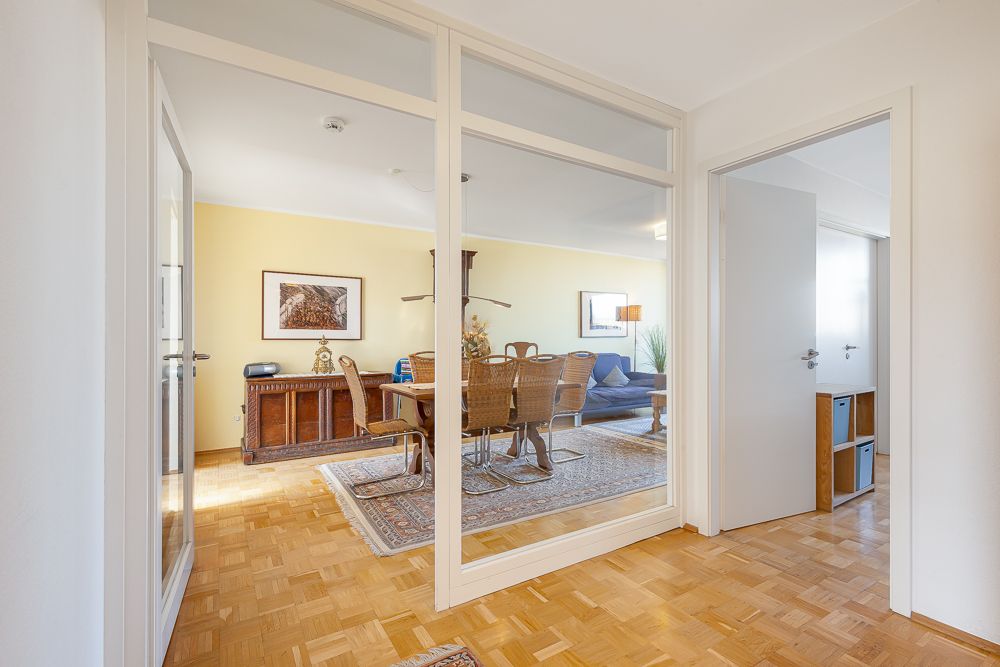 Esszimmer mit Holztisch und Stühlen, daneben ein Wohnzimmer mit Sofa, Glastrennwand im Vordergrund.