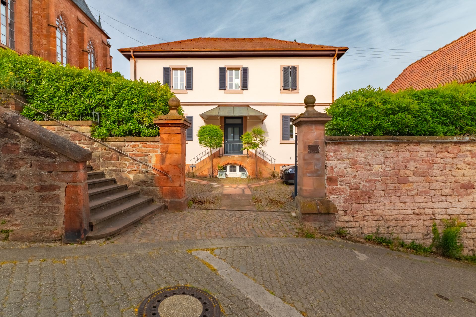 Zweistöckiges Wohnhaus mit rotem Dach und hellen Fassaden, umgeben von Steinmauern und grünen Büschen.