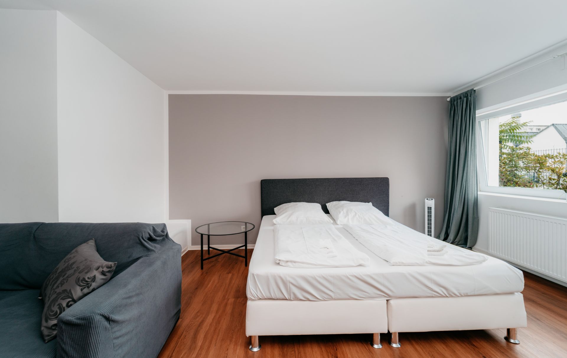 Helles Schlafzimmer mit Doppelbett, grauem Sofa, rundem Tisch und großem Fenster mit Vorhang.