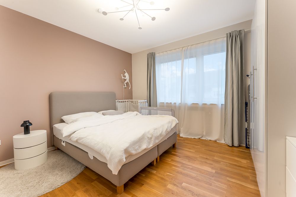 Helles Schlafzimmer mit Doppelbett, Kinderbett, Nachttisch und großem Fenster mit Vorhängen.