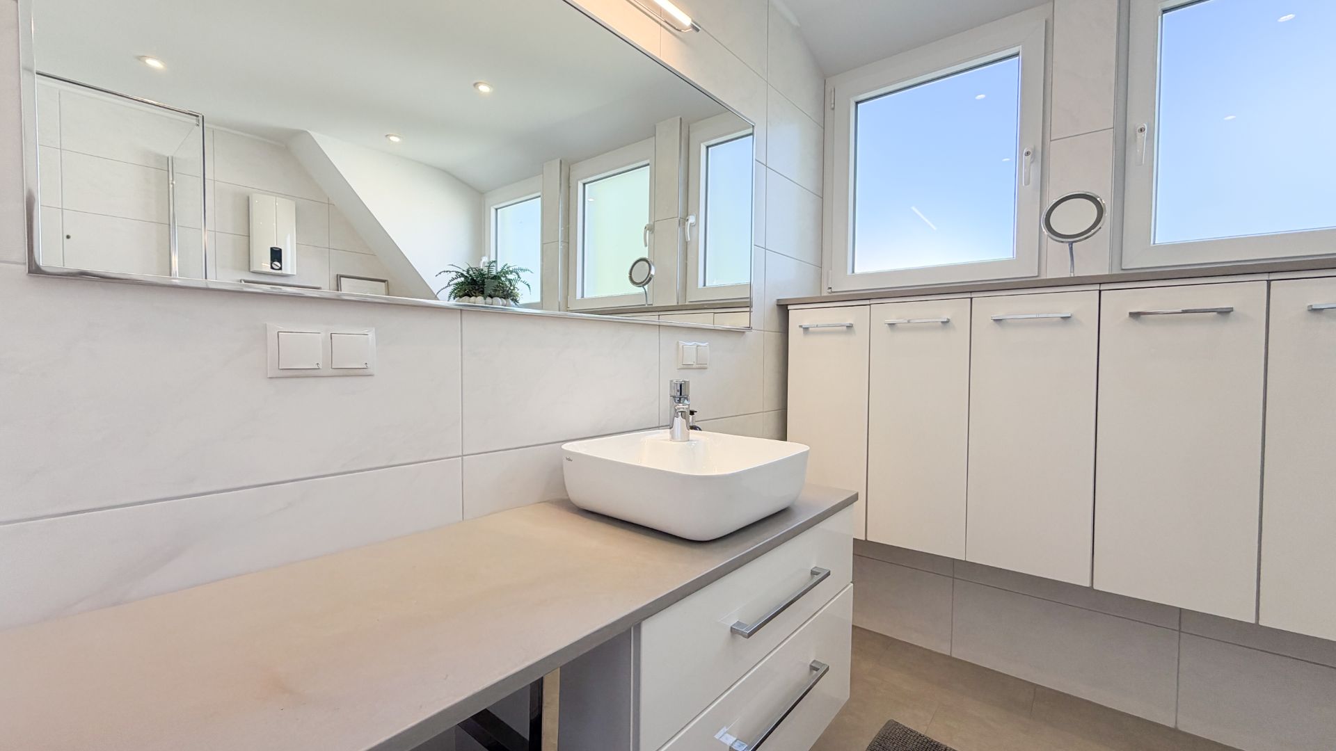 Modernes Badezimmer mit Waschbecken, großem Spiegel und weißen Schränken unter den Fenstern.