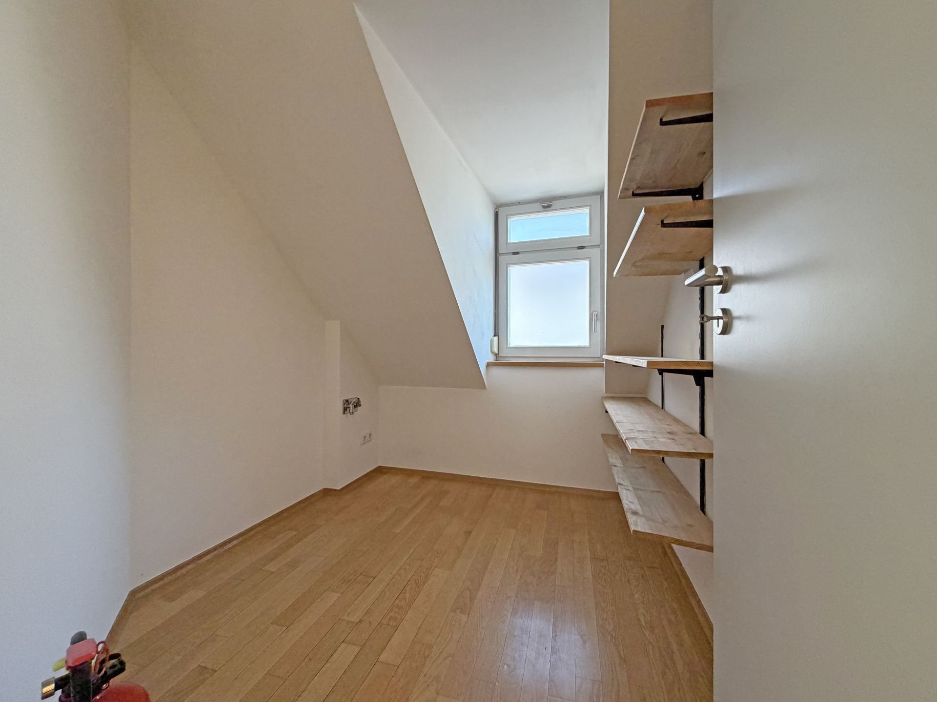 Kleiner Raum mit schräger Decke, Fenster und Wandregalen, Holzboden.
