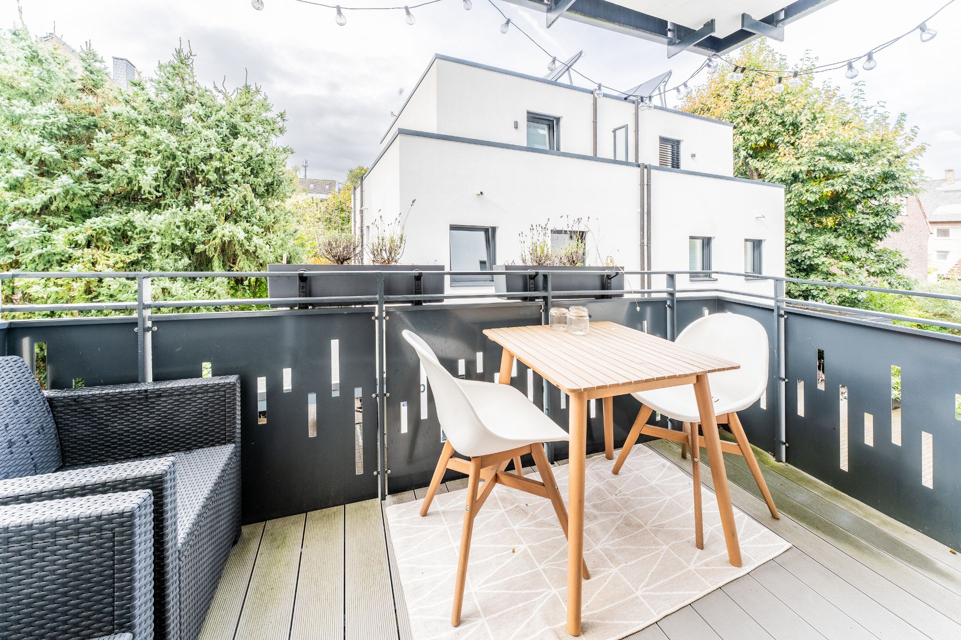 Balkon mit Holztisch, zwei weißen Stühlen und schwarzem Sessel, Blick auf Bäume und modernes Gebäude.