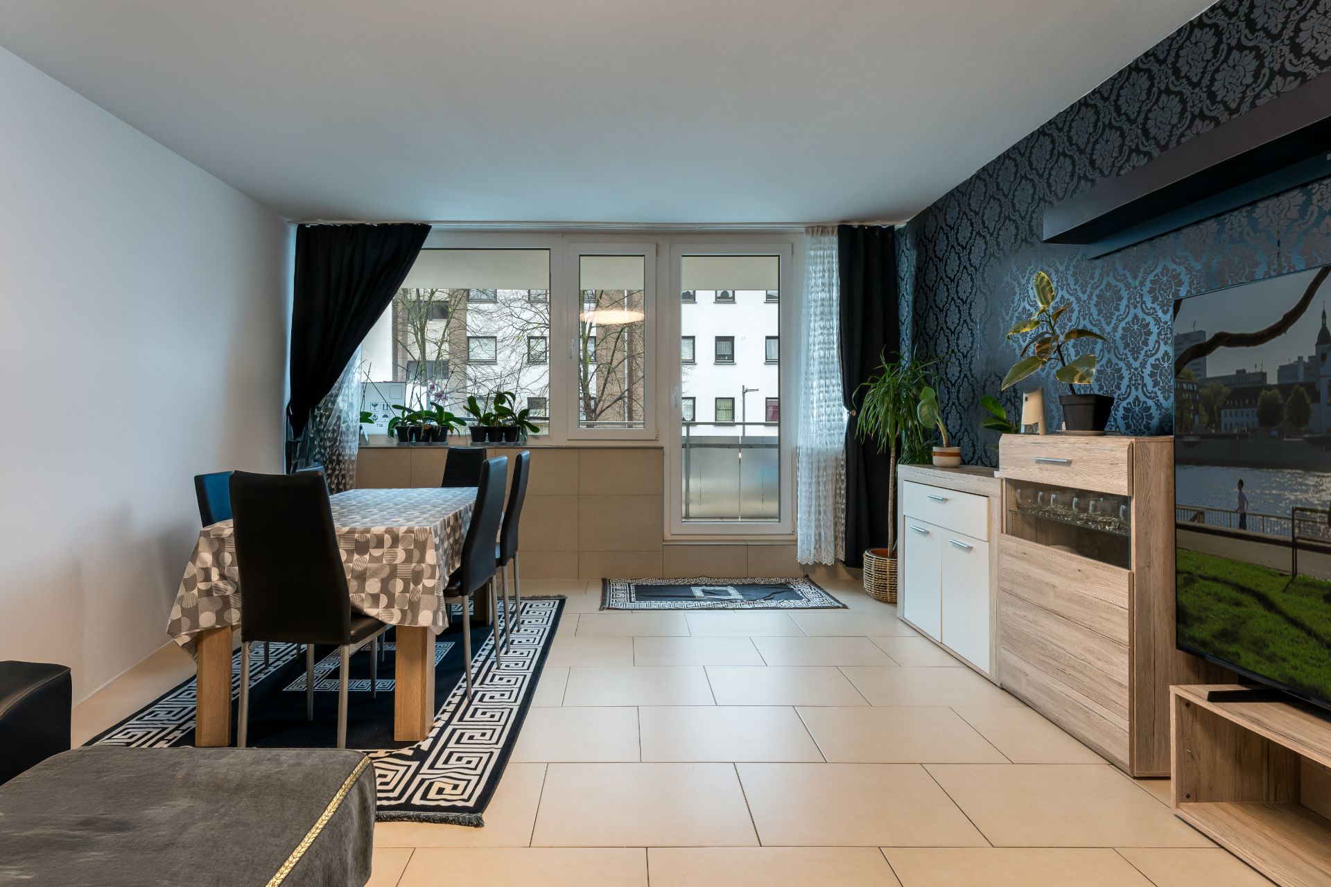 Wohnzimmer mit Esstisch, schwarzen Stühlen, Sideboard, Pflanzen und großem Fenster.