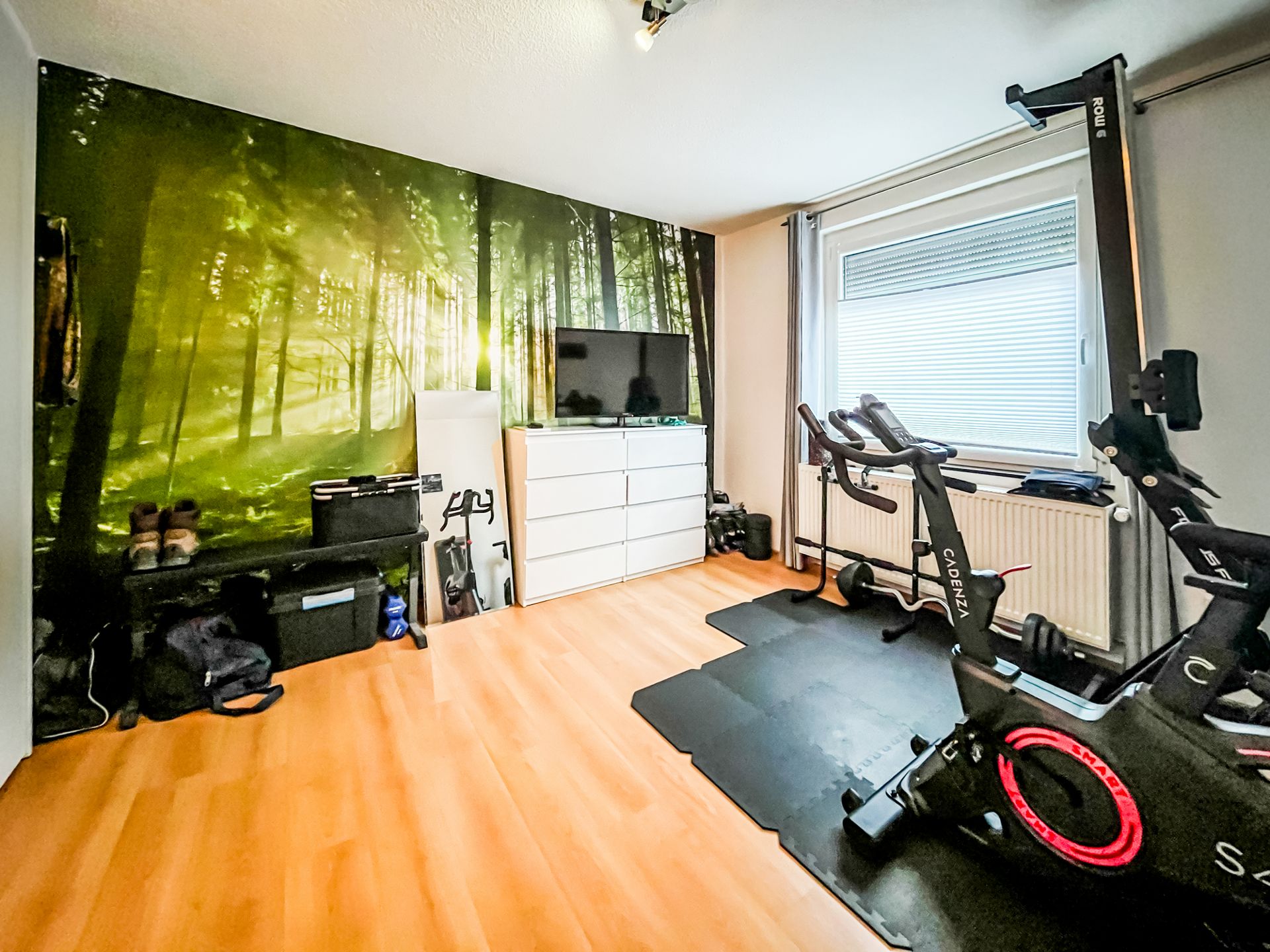 Heller Fitnessraum mit Heimtrainer, Fernseher, Fenster und Wandbild von einem Wald.