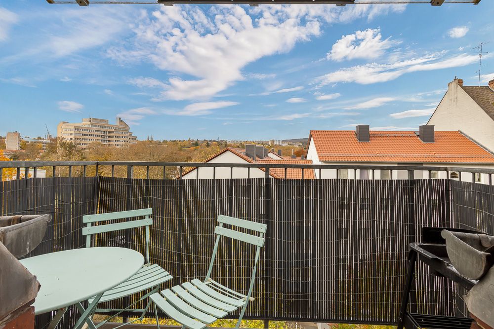 Kleiner Balkon mit zwei Stühlen, Tisch und Blick auf Nachbarhäuser unter blauem Himmel.