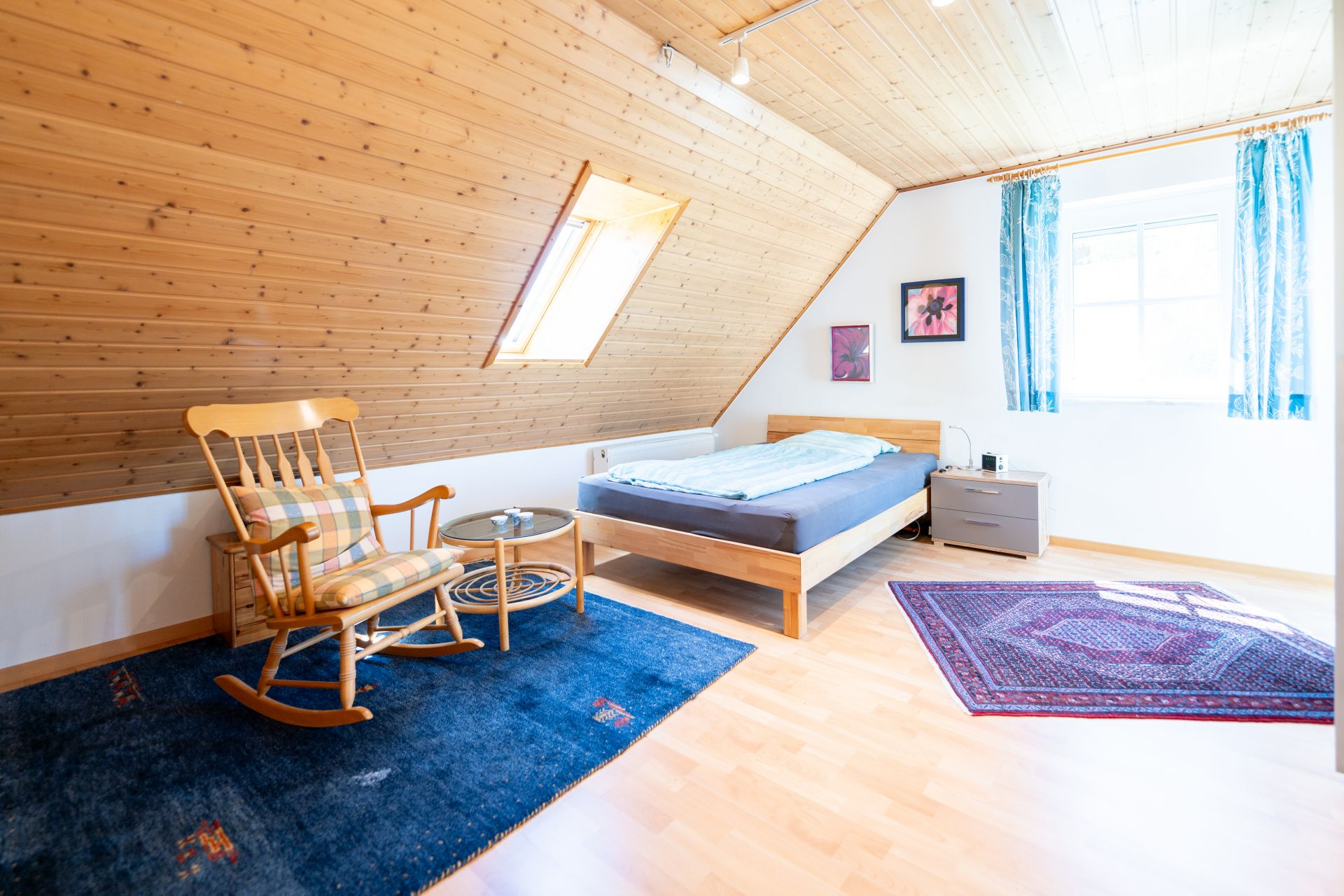 Helles Schlafzimmer mit Dachschräge, Holzdecke, Bett, Schaukelstuhl und Teppichen.