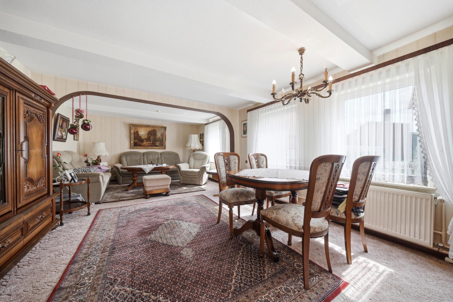 Wohnzimmer mit Teppich, Esstisch, Stühlen, Sofa und großen Fenstern mit Gardinen.