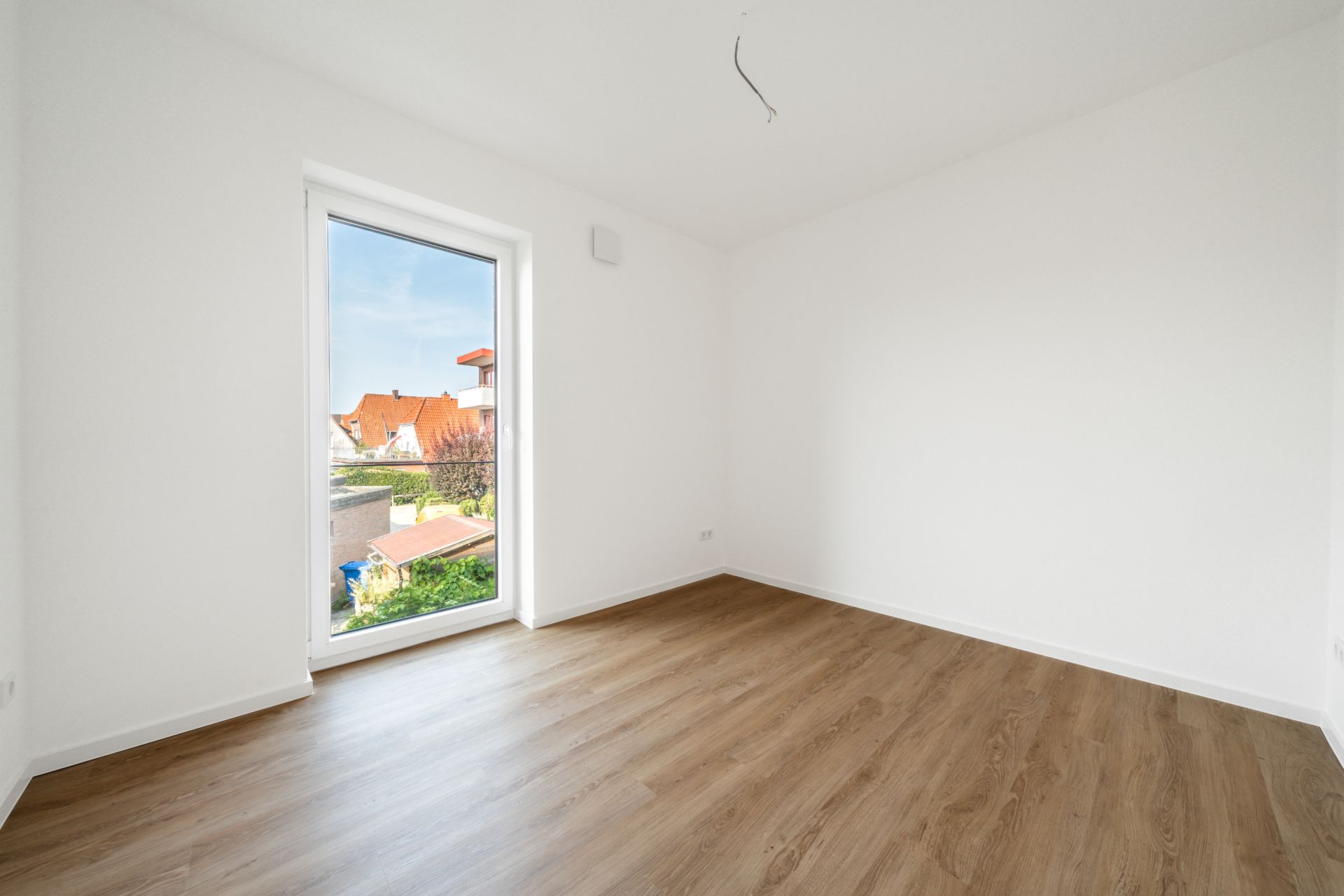Leeres Zimmer mit weißer Wand, großem Fenster und Holzboden.