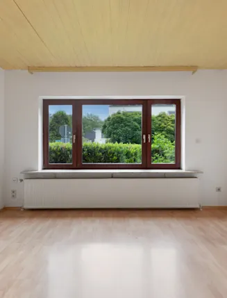 Leerer Raum mit hellem Holzboden, großem Fenster und Blick auf grüne Büsche draußen.