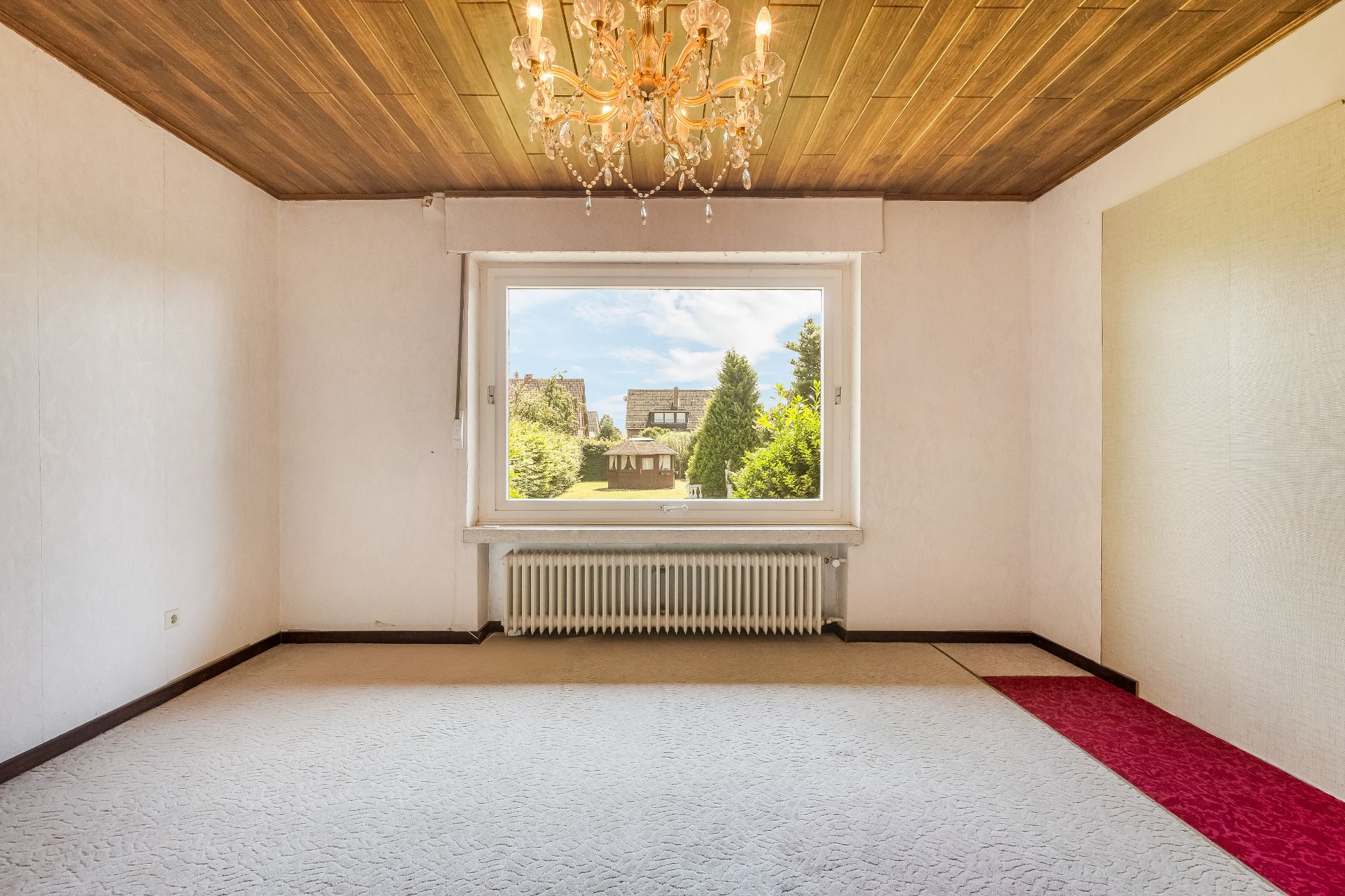 Leerer Raum mit Holzbalkendecke, Kronleuchter und großem Fenster mit Blick ins Grüne.