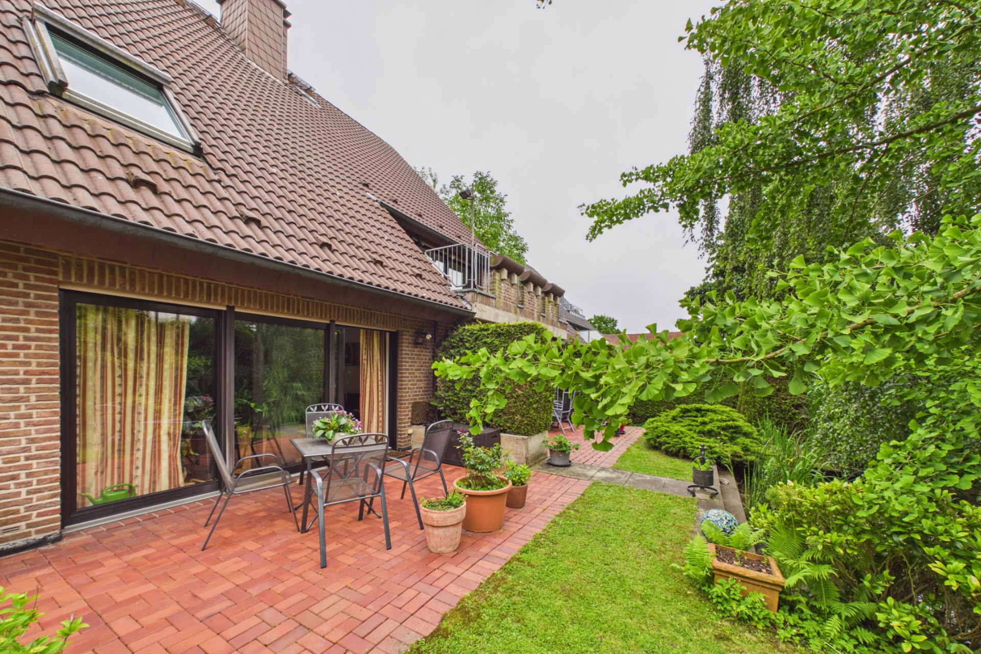 Terrasse mit Tisch, Stühlen und Pflanzen an einem Backsteinhaus, daneben ein Garten mit viel Grün.