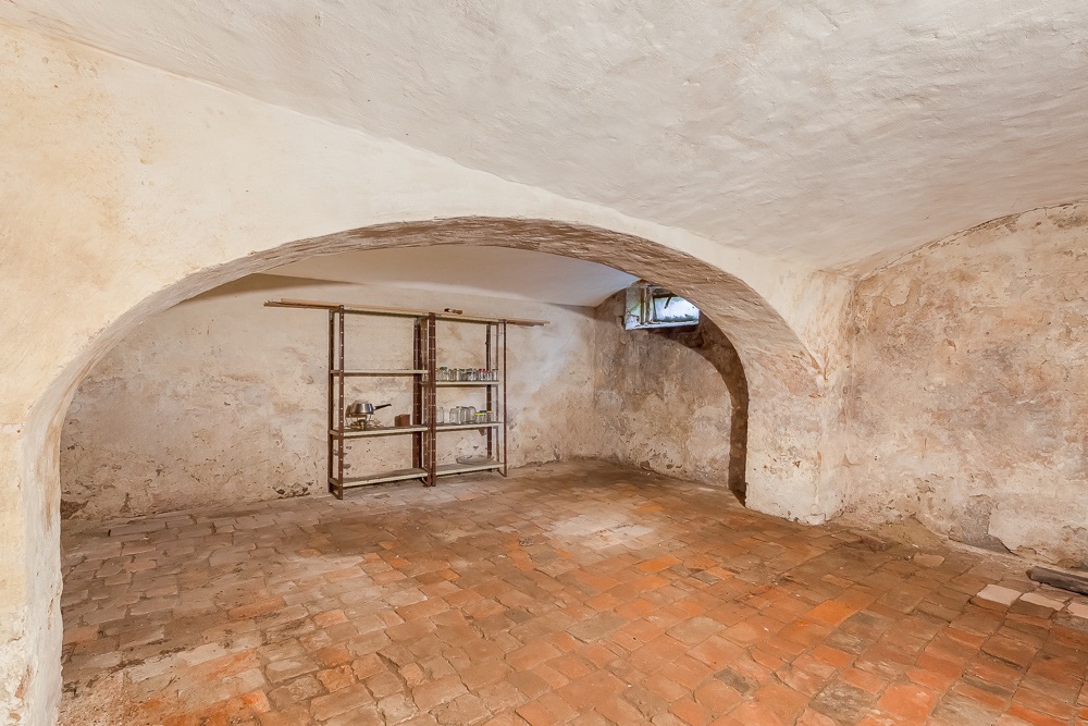 Alter Kellerraum mit Gewölbedecke, Steinboden und Metallregal an der Wand.