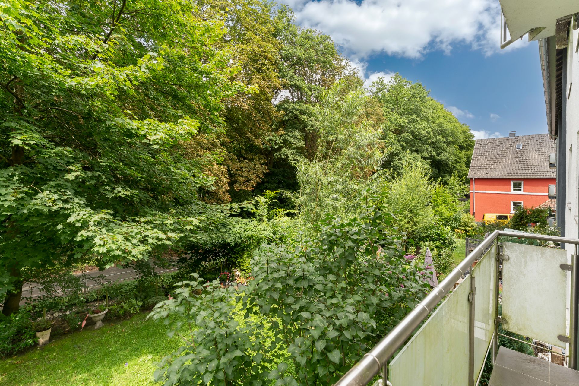 Blick von einem Balkon auf einen grünen Garten mit Bäumen und Nachbarhaus im Hintergrund.