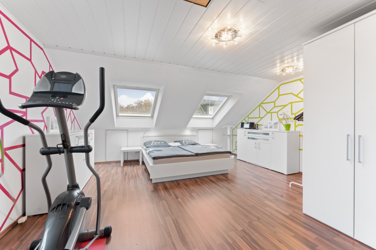 Modernes Schlafzimmer mit Dachfenstern, Bett, Fitnessgerät und weißen Möbeln auf Holzfußboden.