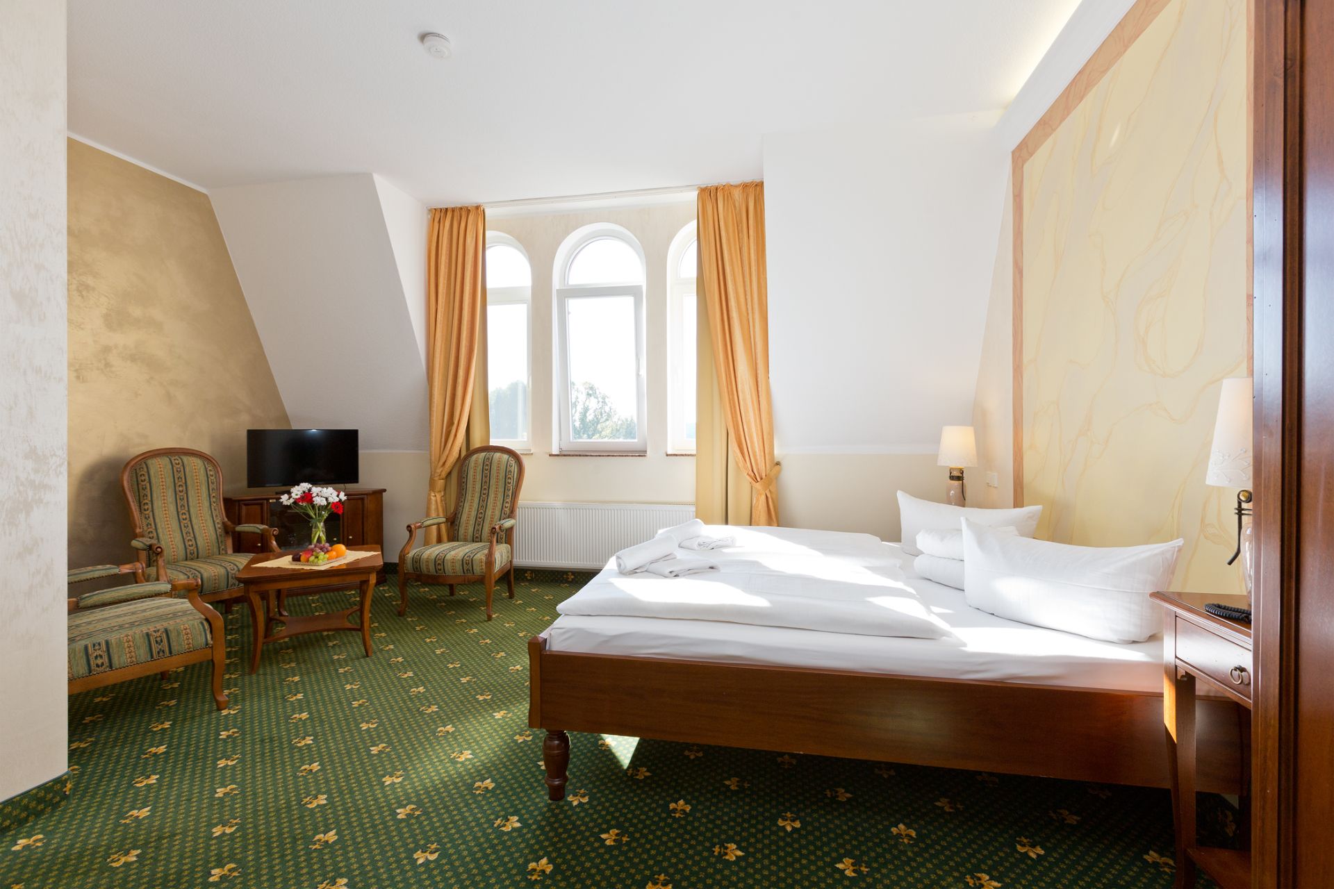 Helles Schlafzimmer mit Doppelbett, zwei Sesseln, kleinem Tisch, Fernseher und drei Fenstern mit gelben Vorhängen.
