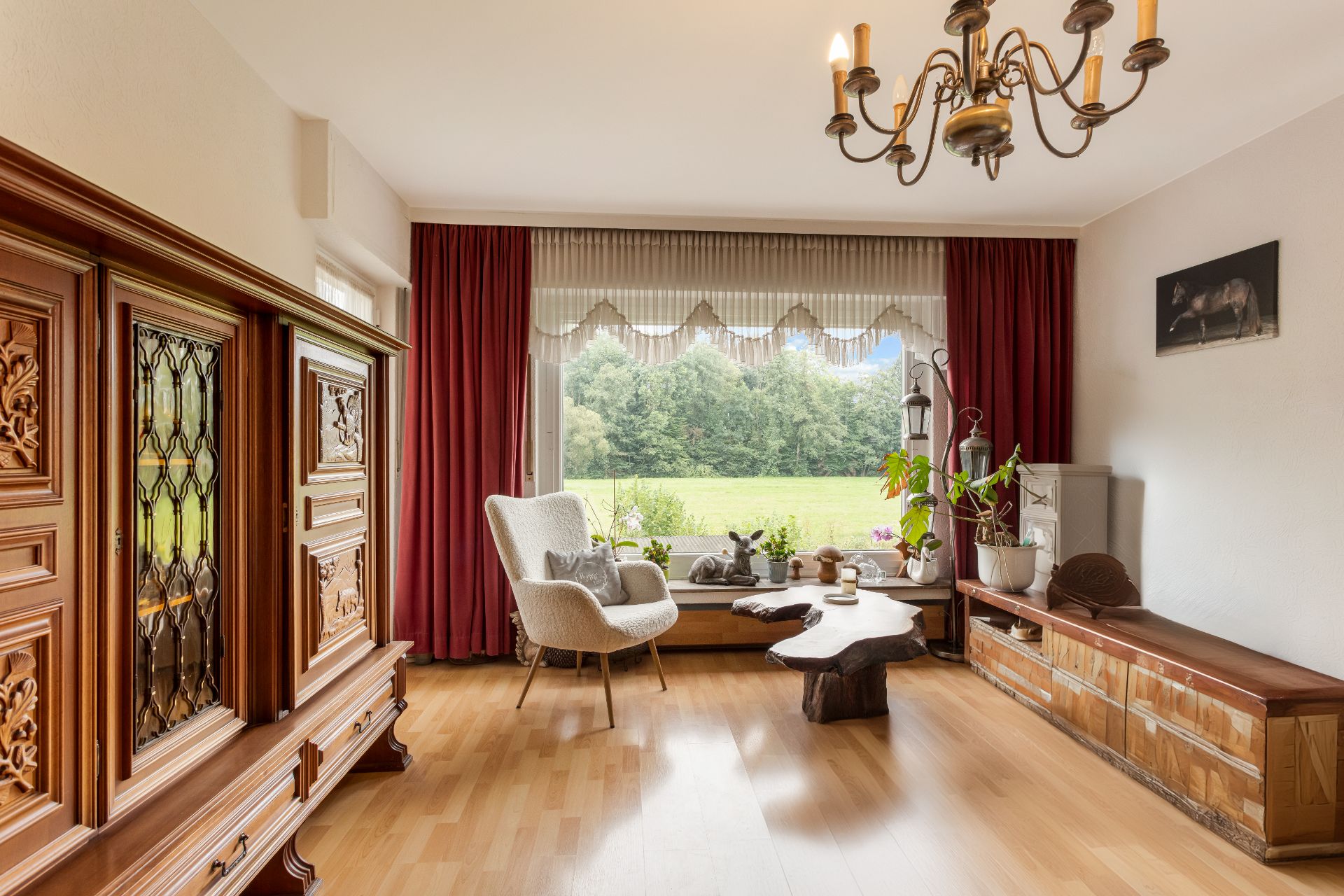 Wohnzimmer mit Holzschrank, Sessel, Holztisch und großem Fenster mit Blick ins Grüne.