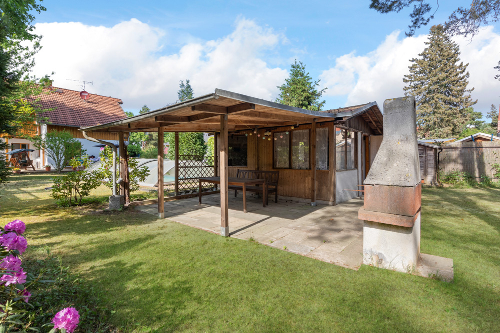 Überdachte Terrasse mit Holztisch und Garten im Grünen, daneben ein gemauerter Grill.