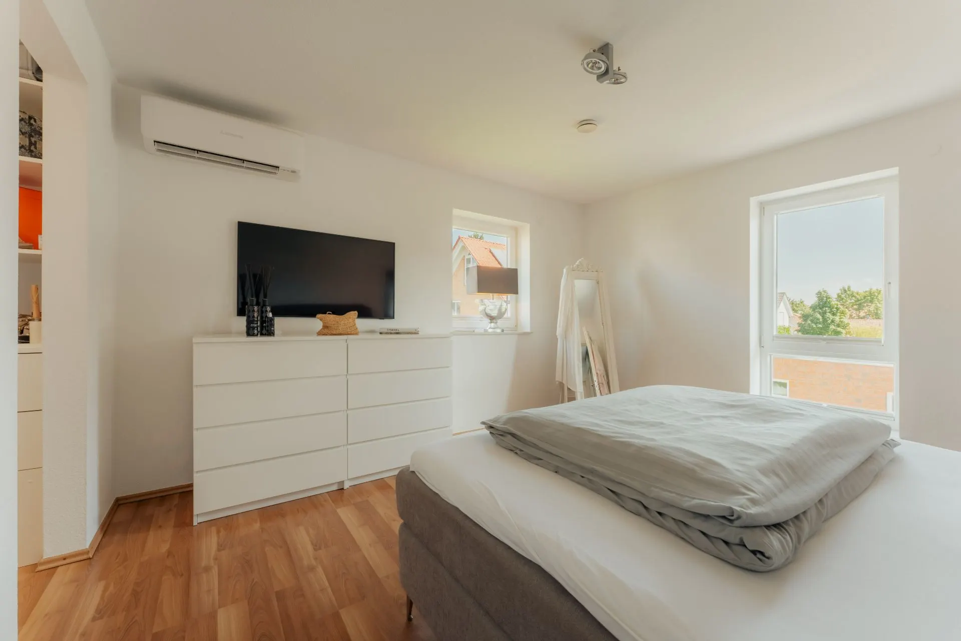 Helles Schlafzimmer mit Doppelbett, Kommode, Fernseher und großen Fenstern.